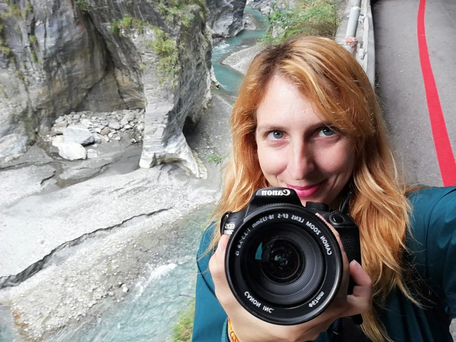 A woman with reddish hair taking a selfie with a Canon camera in front of a rocky canyon with a river below.