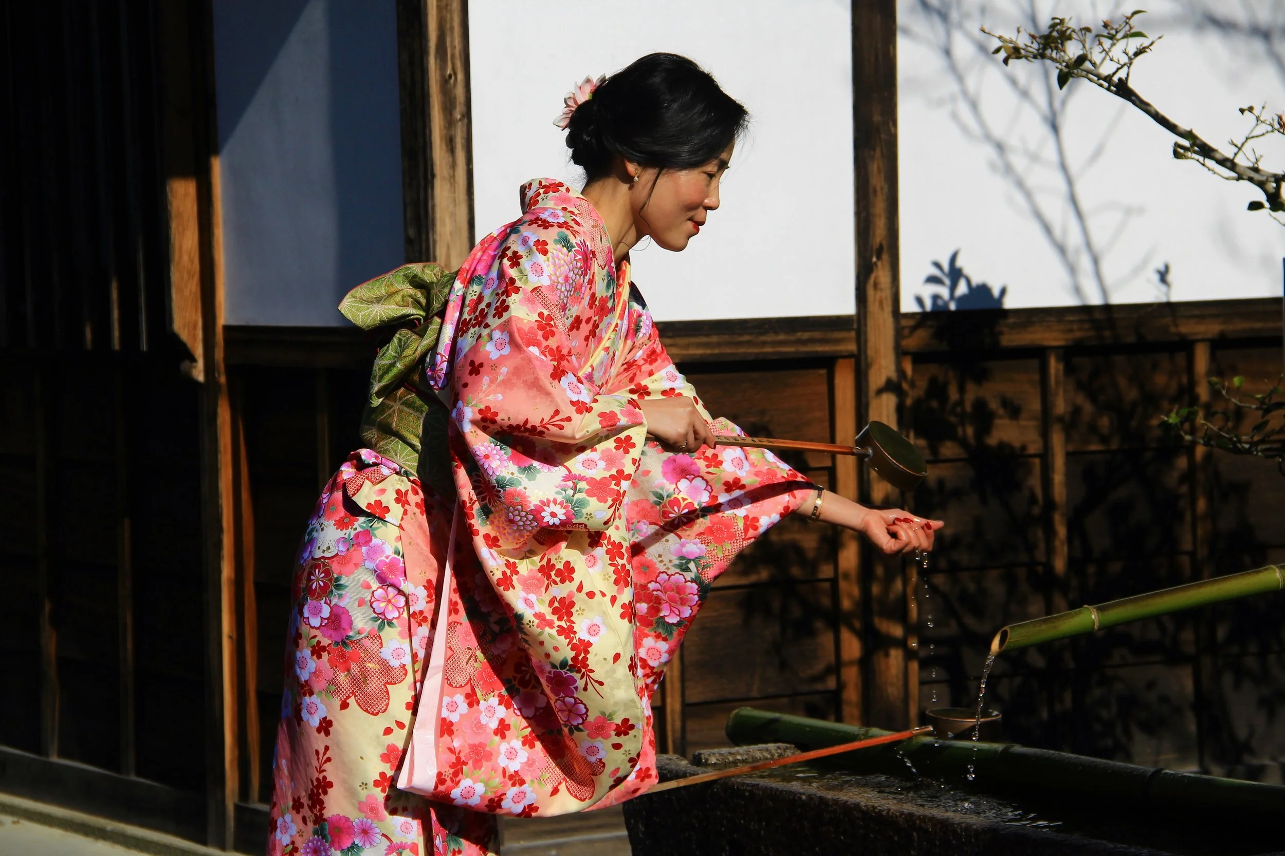 A woman dressed in a pink floral kimono waters her hand with a bamboo ladle at a traditional Japanese water basin outdoors.