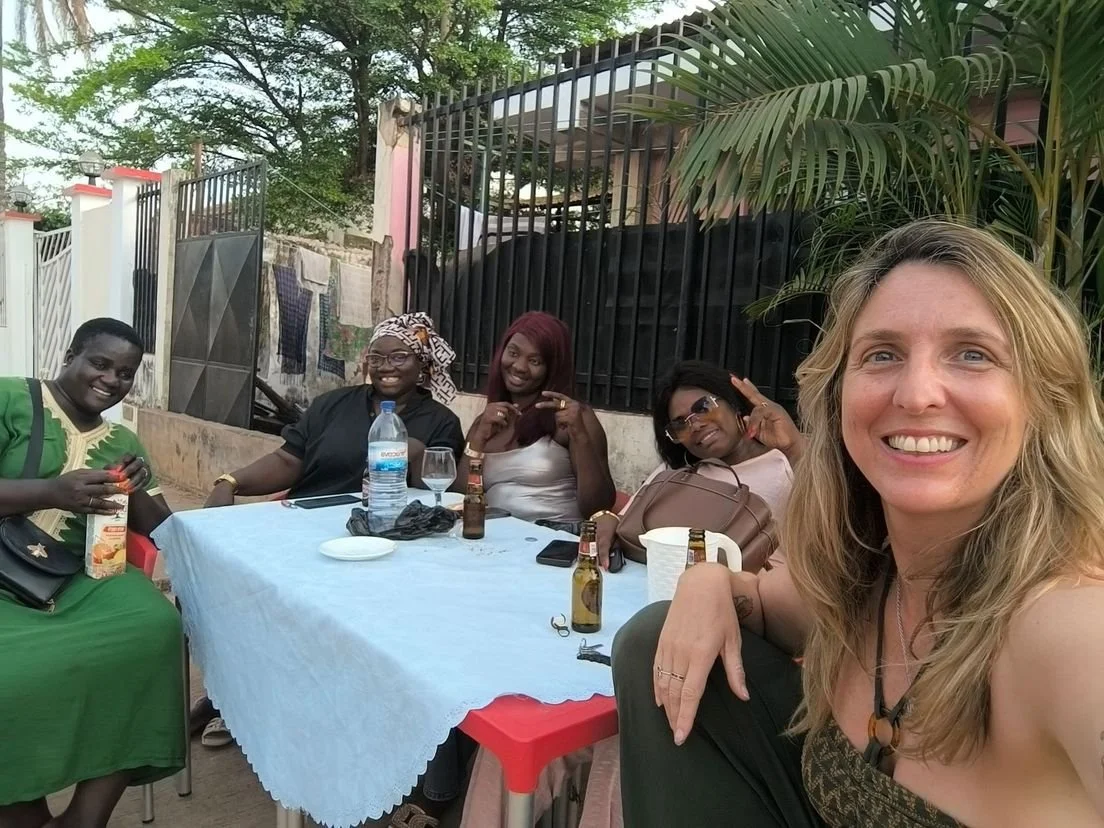 Group of five women sitting at an outdoor table, smiling. The woman taking the selfie has blonde hair and is wearing a black top. The table has drinks, a water bottle, and personal belongings. There is a black metal fence and greenery in the backgrou