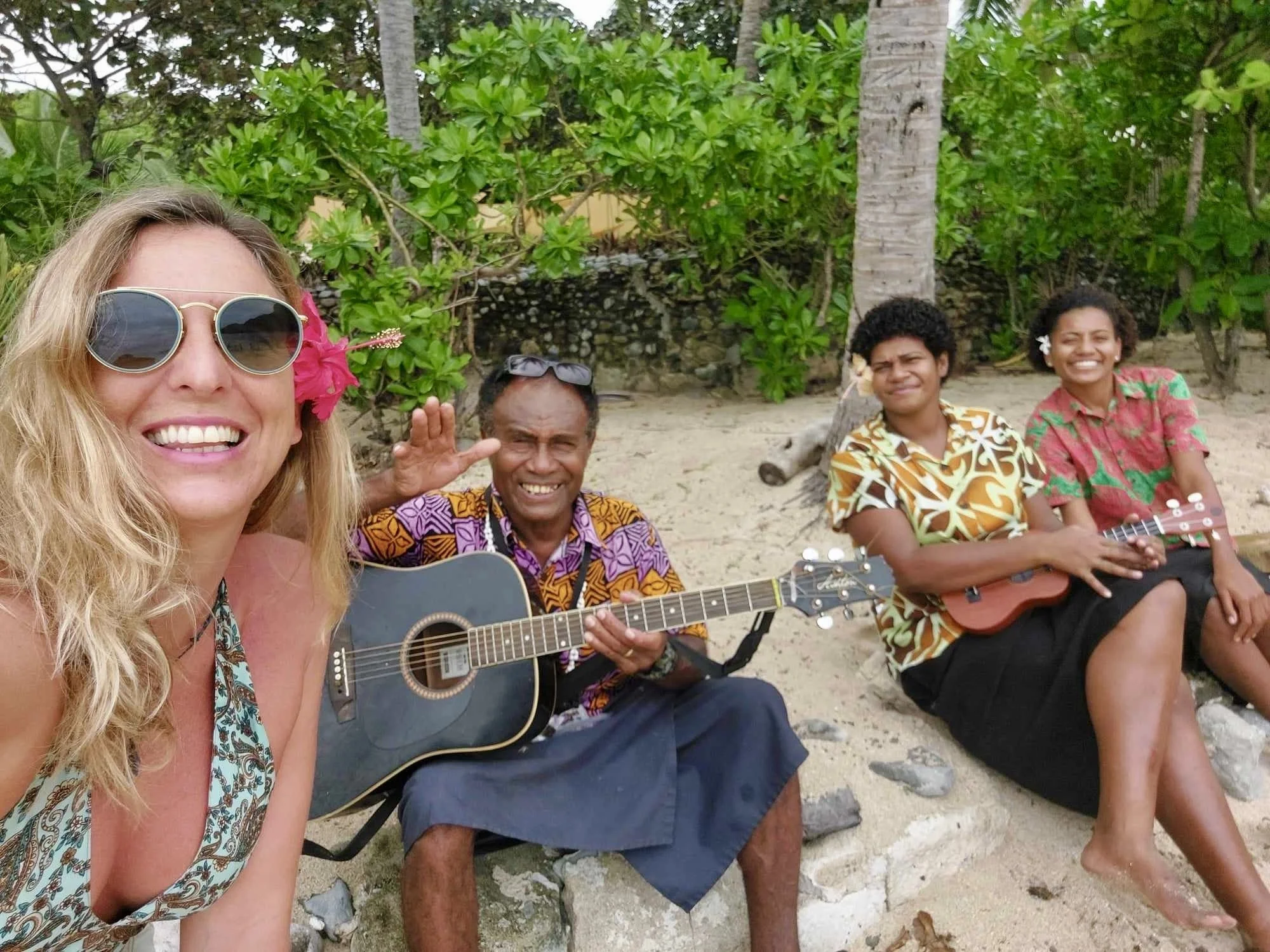 Group of four people sitting on rocks and playing guitars on a sandy beach with green foliage and palm trees in the background. One woman is taking a selfie, wearing sunglasses and a flower in her hair, smiling; a man next to her is holding a guitar 