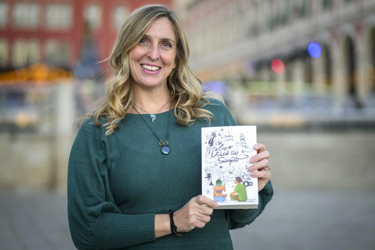 Woman with blonde hair smiling outdoors, holding a book with an illustrated cover featuring two children sitting and talking, cityscape background.