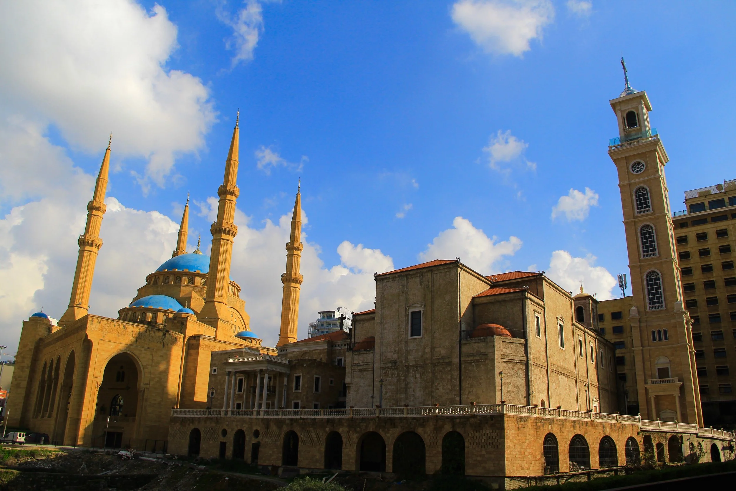 A mosque with a large central blue dome and four minarets, along with a tall clock tower against a partly cloudy sky.