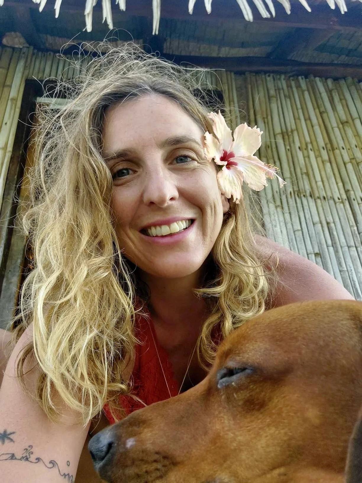 A woman with curly blonde hair and a pink flower in her hair, smiling at the camera, with a brown dog resting its head near her. The background features bamboo walls and a wooden roof.