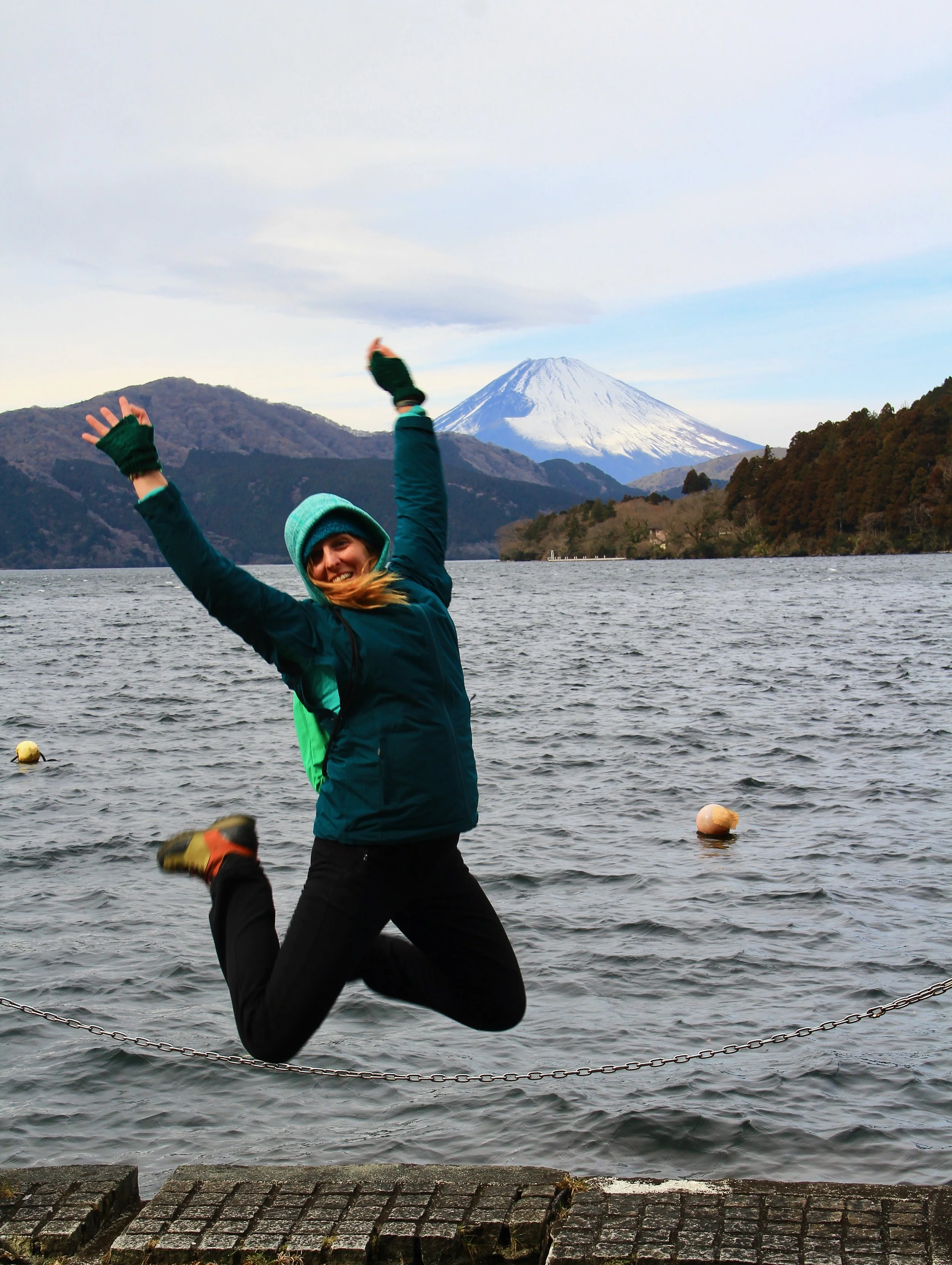 Woman in outdoor winter gear leaping over a chain barrier by a lake with mountains and snow-capped volcano in the background.