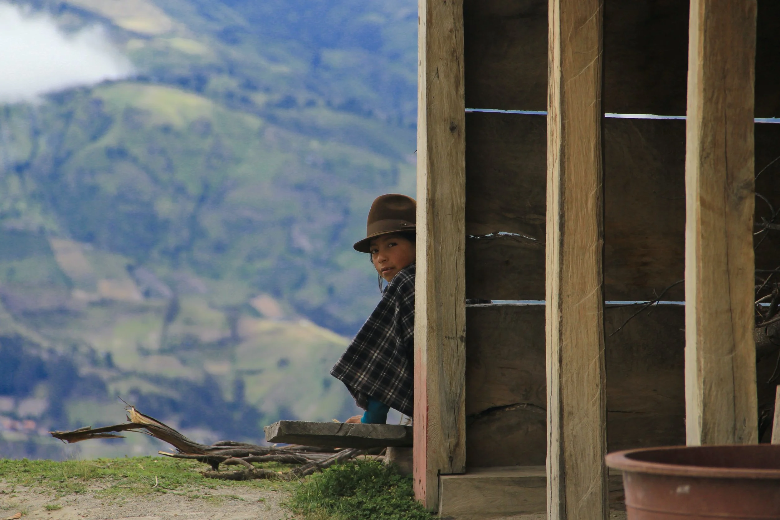 A young child wearing a brown hat and a checkered shawl peeks out from behind a wooden hut with mountains in the background.