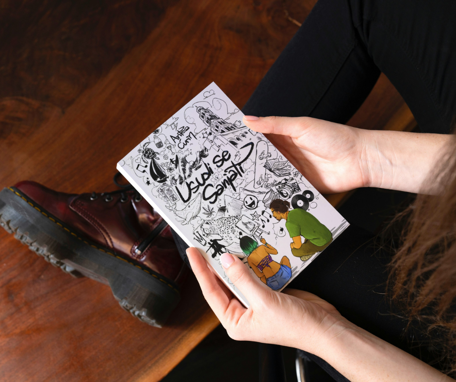 Person wearing brown boots sitting on wooden floor, holding a comic book with a black-and-white and colorful illustration of two characters reading.
