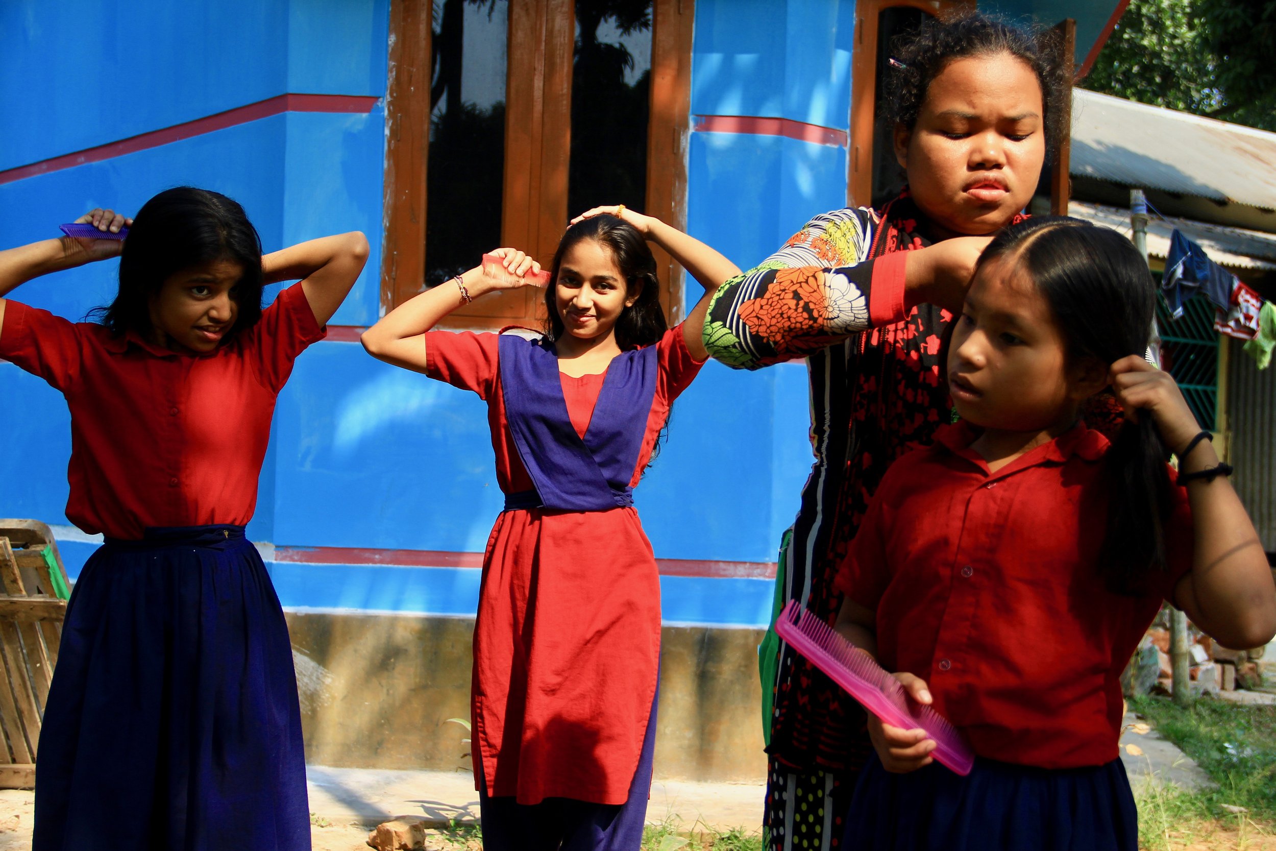 Four young girls in colorful dresses standing outdoors near a blue building, one girl appears to be styling her hair, another holding a pink comb, and a woman is assisting a girl with her hair, all involved in hair styling activities.