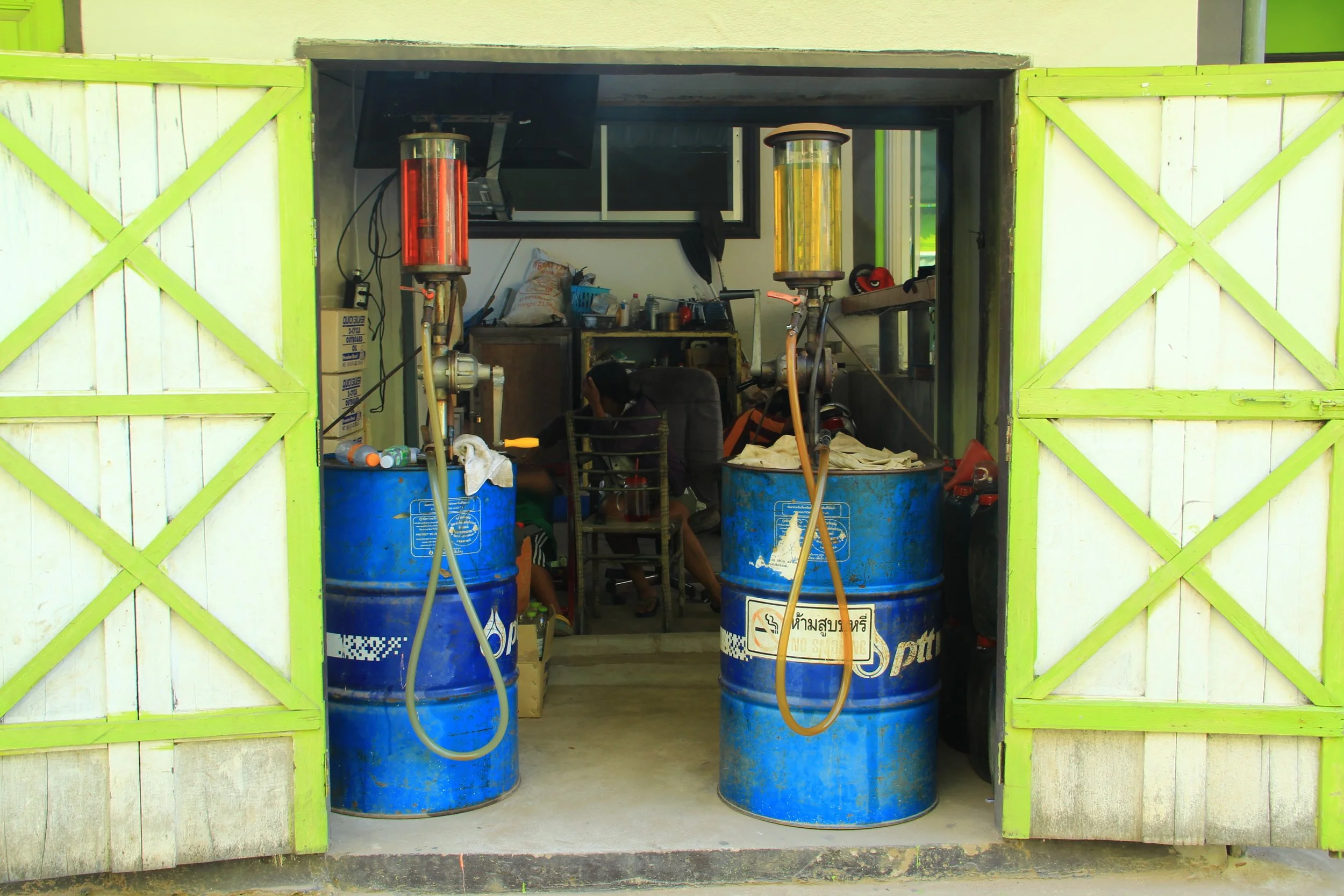 Inside a small workshop with two large blue drums, each with a colored flame tube on top, used for fueling or heating. There are various tools and boxes inside, and a person is sitting and working at a table.
