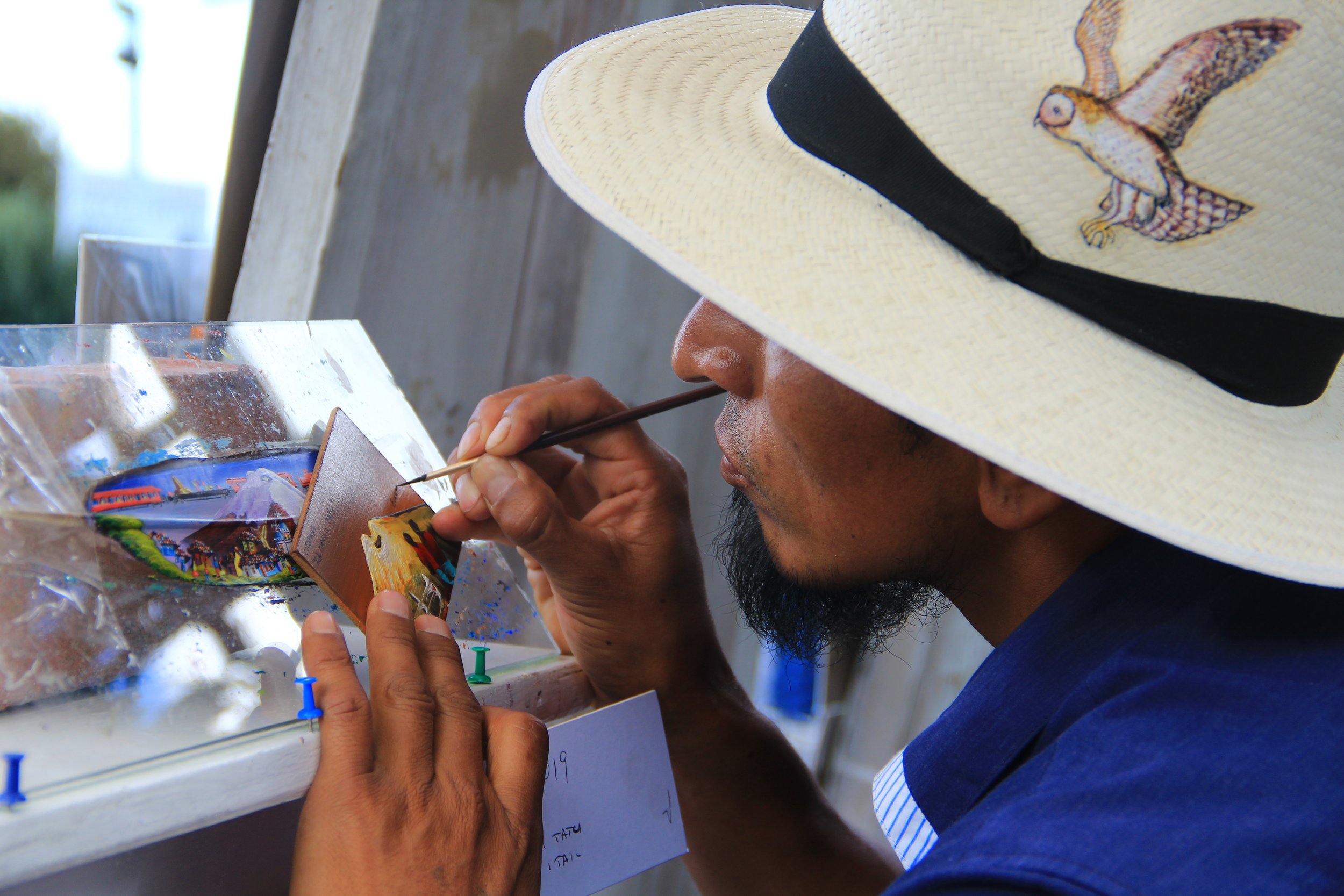 A man with a painted straw hat featuring a bird design is painting a small, colorful piece of artwork on a wooden or canvas surface near a window, with a glass of water and paint supplies nearby.