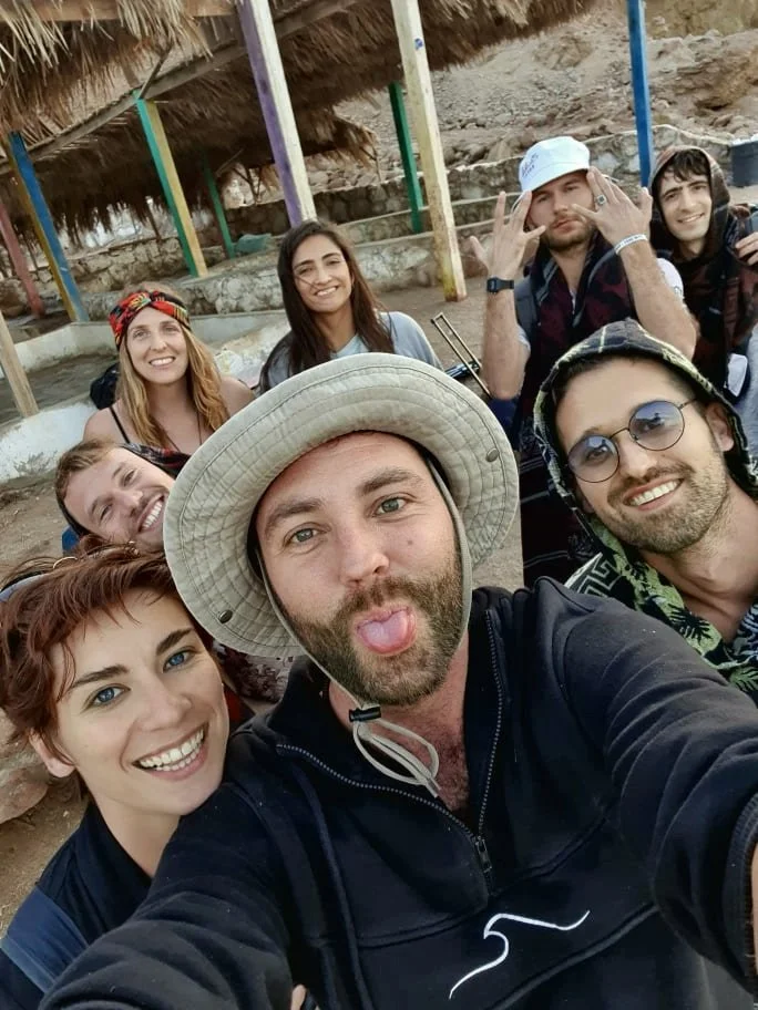 Group of eight friends taking a selfie outdoors, some smiling and one sticking out tongue, with colorful wooden posts and thatched roof structures in the background.