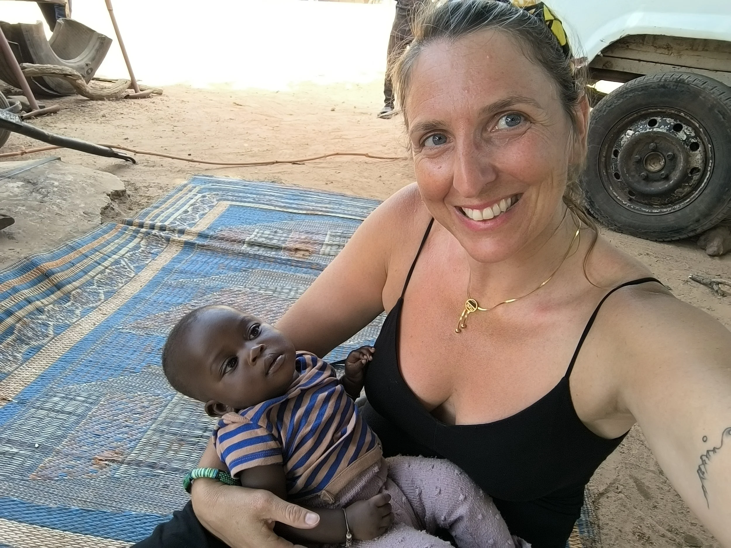 A woman with light skin and blonde hair taking a selfie while holding a young child with dark skin, in an outdoor setting with dirt ground, a colorful mat, a vehicle tire, and some tools visible in the background.