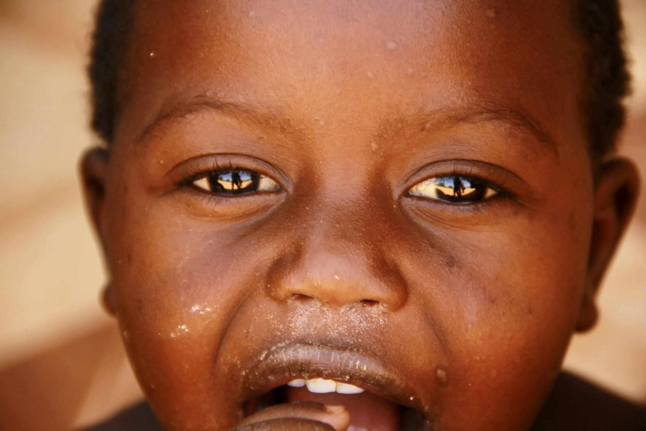 Close-up of a young child's face with a curious expression, showing dark skin, shorthair, and reflections of trees and sky in the eyes.