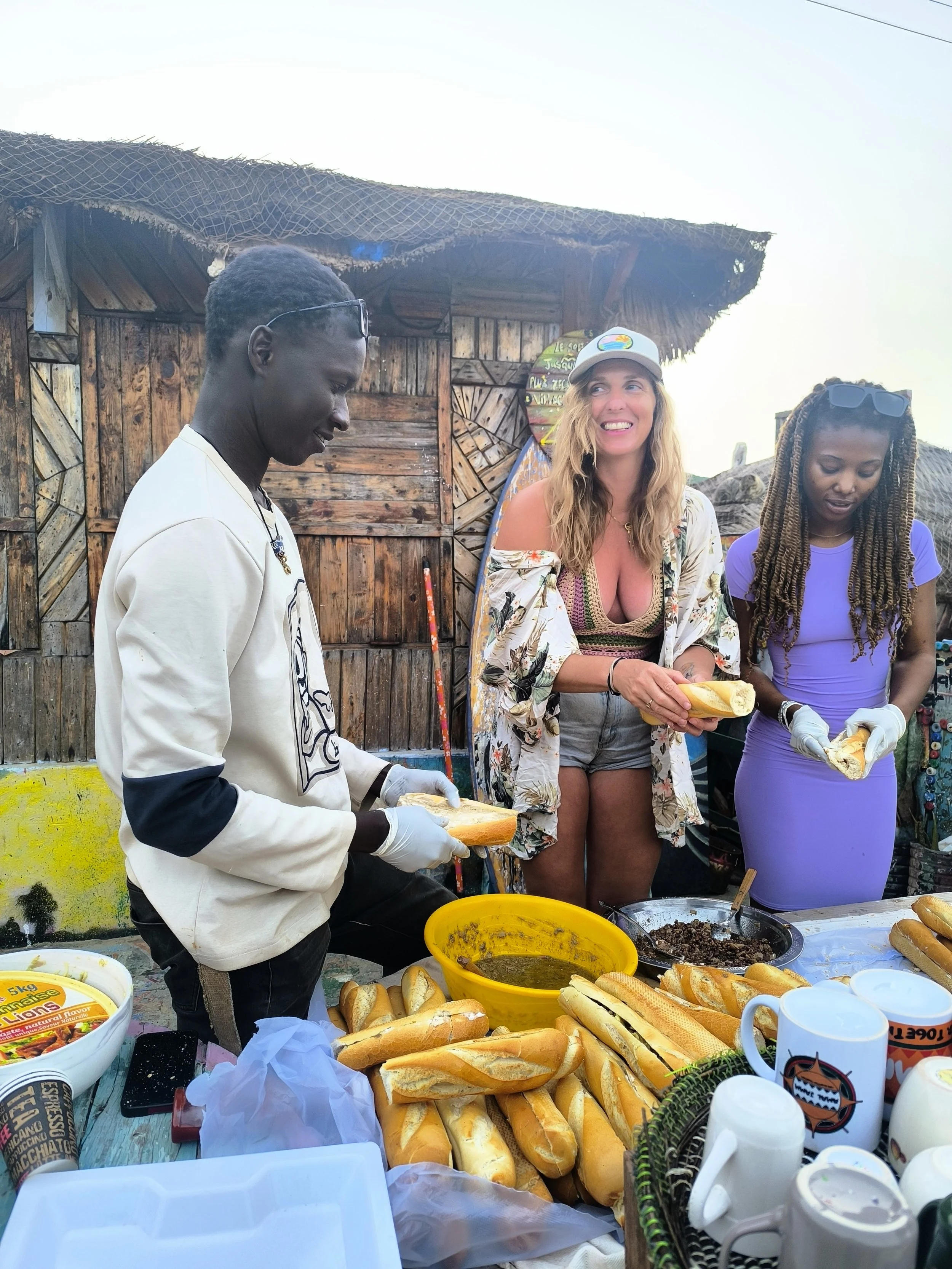 Three women preparing sandwiches outdoors in front of a wooden shed, with a table filled with baguettes, condiments, and mugs.