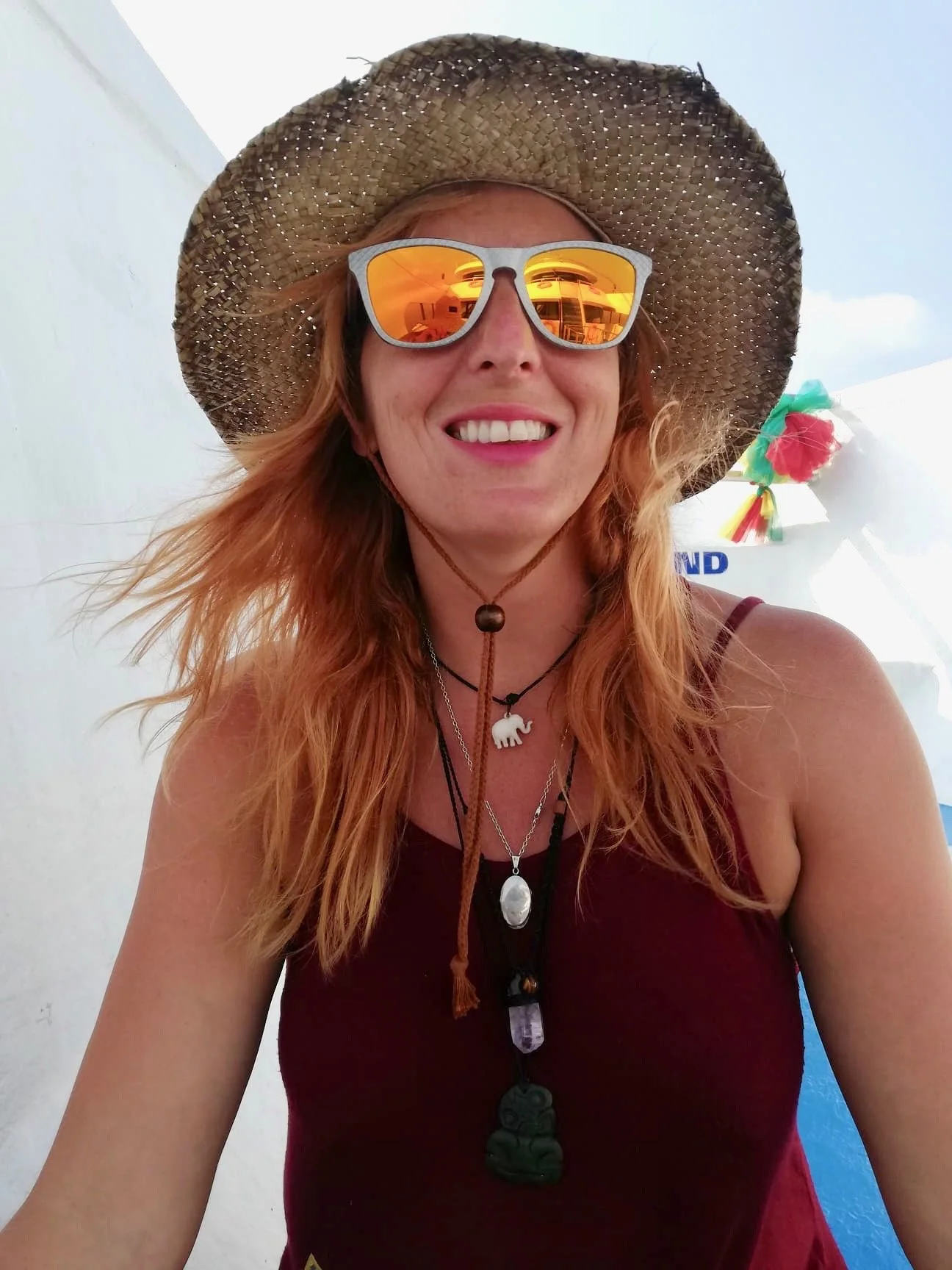 A smiling woman with red hair wearing a straw hat, reflective sunglasses, a maroon tank top, and layered necklaces, outdoors on a sunny day.