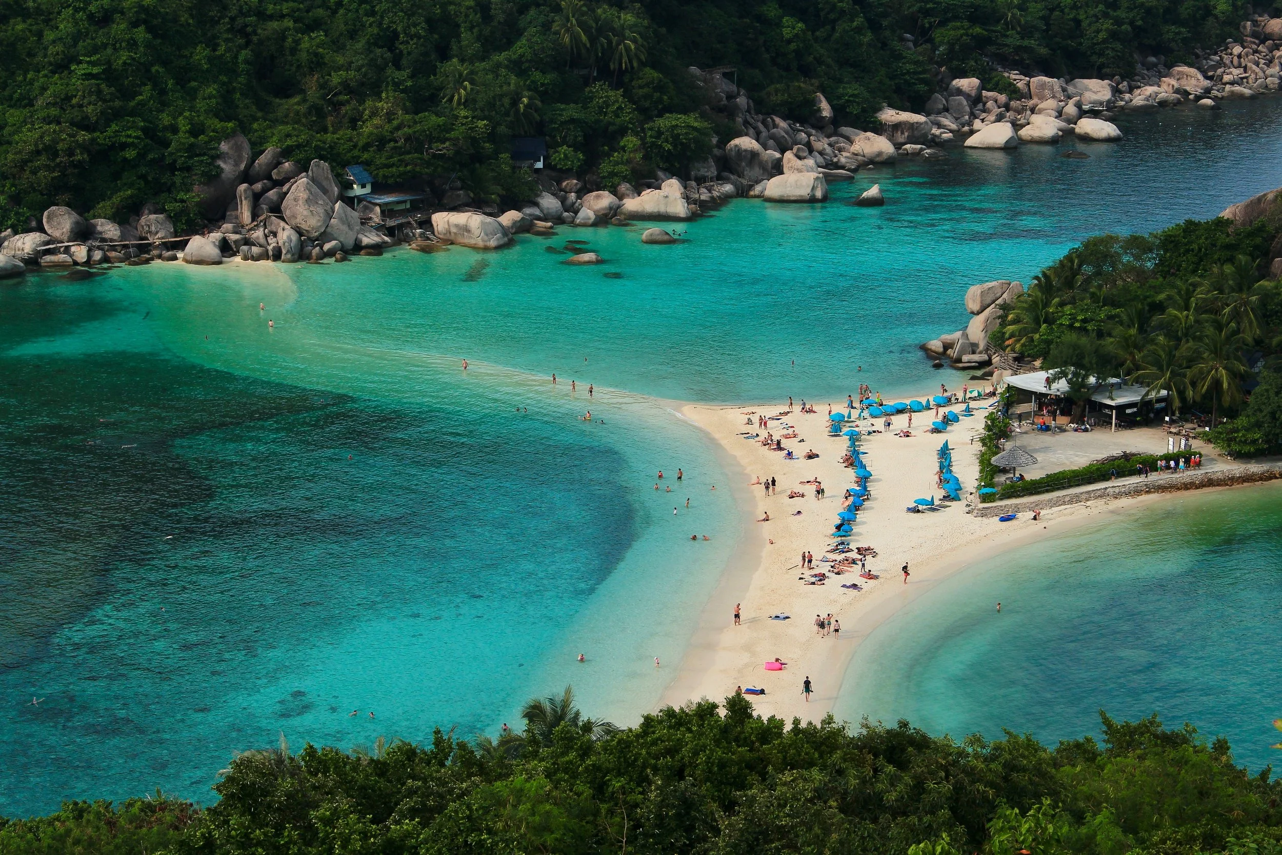 Tropical beach with white sand, turquoise waters, and green foliage in the background, with people lounging and swimming.