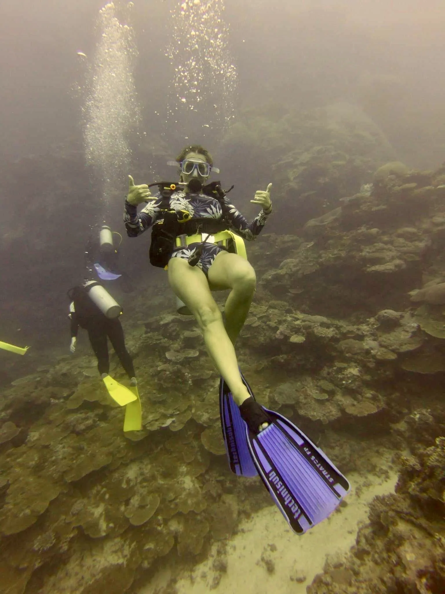 A scuba diver underwater giving a thumbs-up while sitting on a yellow underwater seat, with another diver in the background exploring a coral reef.