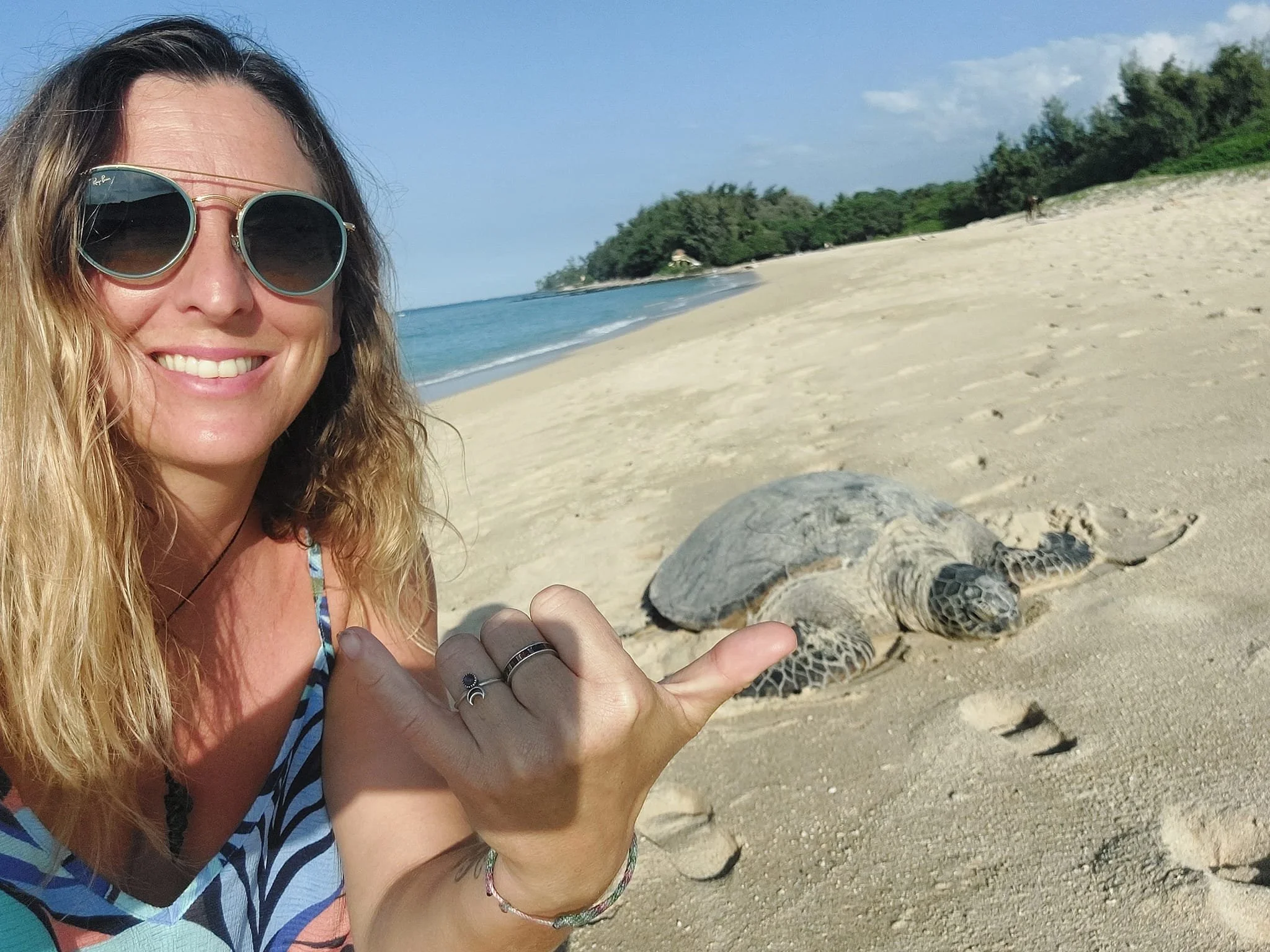 A woman with long blonde hair wearing sunglasses and a patterned swimsuit taking a selfie on a sandy beach with a sea turtle nearby.