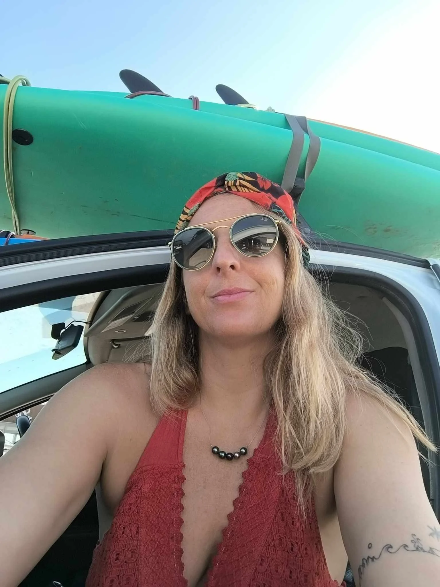 A woman with blonde hair wearing sunglasses, a red sleeveless top, and a red bandana on her head, taking a selfie in front of a vehicle with surfboards on top, at a beach or outdoor location.