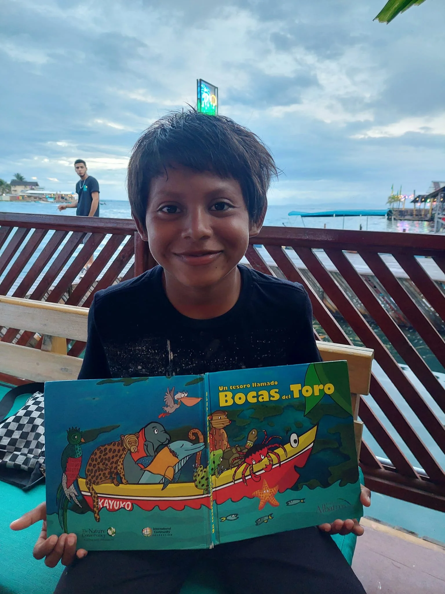 Smiling boy holding a colorful children's book titled 'Un tesoro llamado Bocas del Toro' at a waterfront restaurant with a view of the ocean, boats, and a cloudy sky in the background.