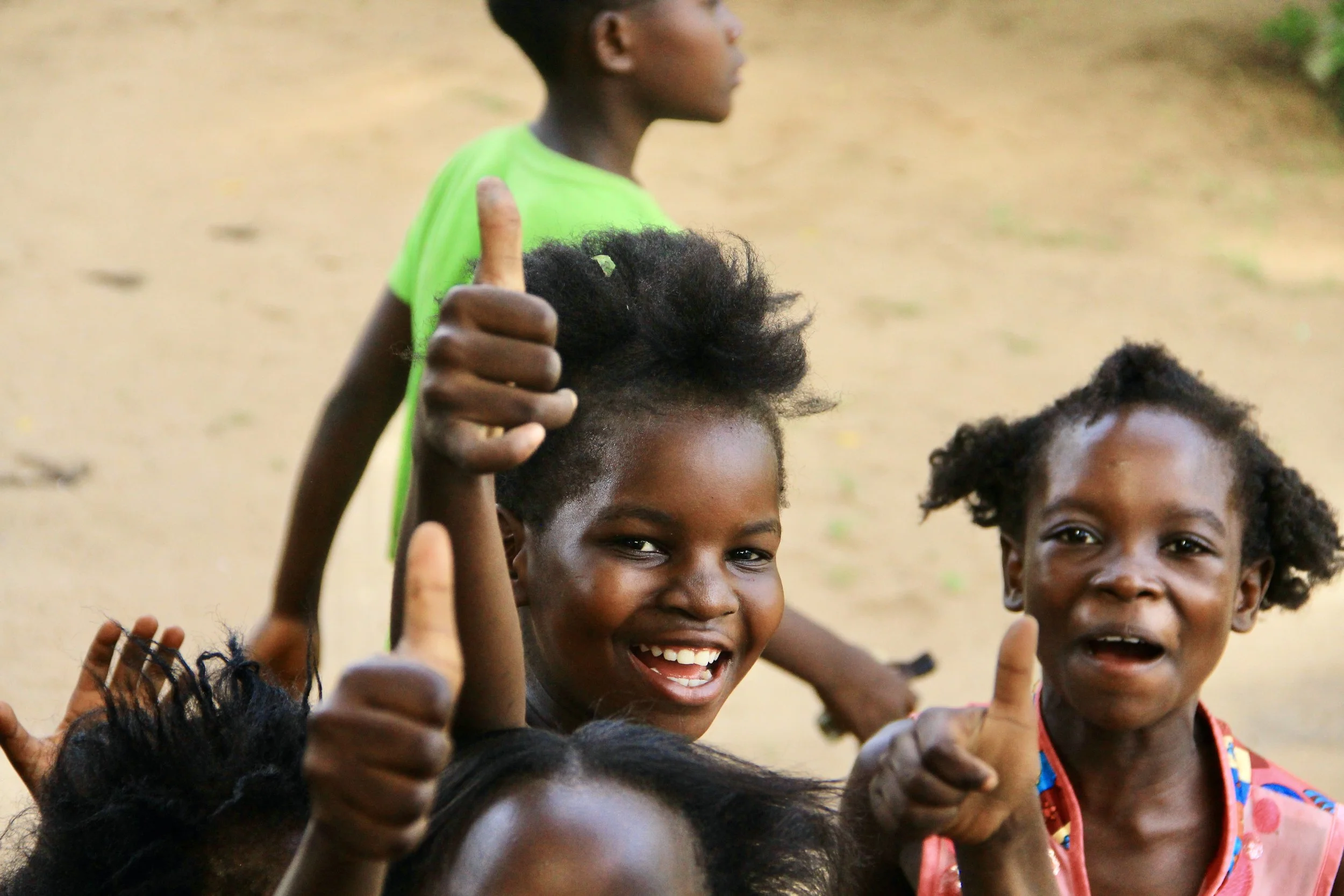 Smiling children giving thumbs up, with a girl in the foreground and others in the background on a dirt ground.