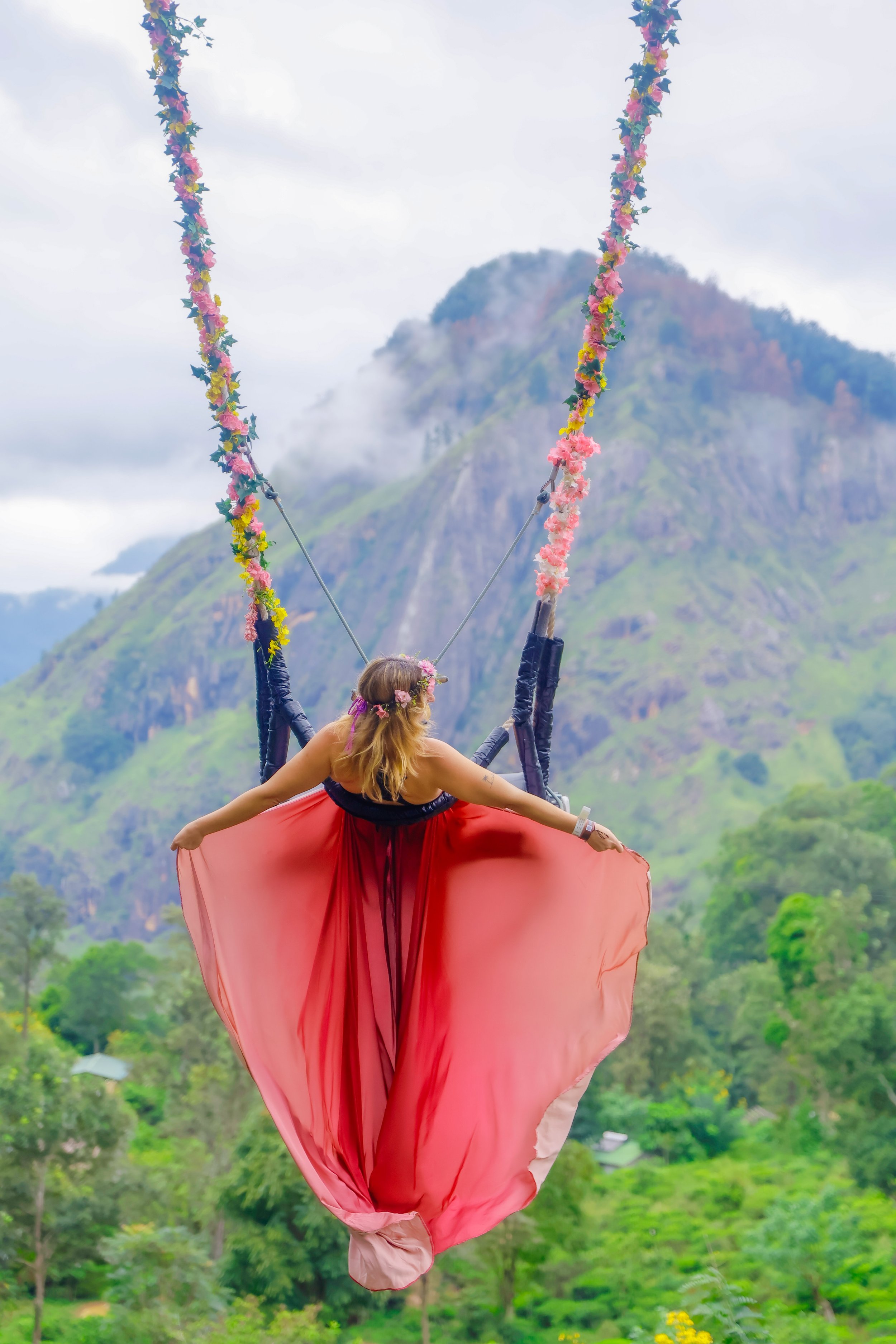 A woman in a pink flowing dress swinging on a floral swing with a mountain and green landscape in the background.