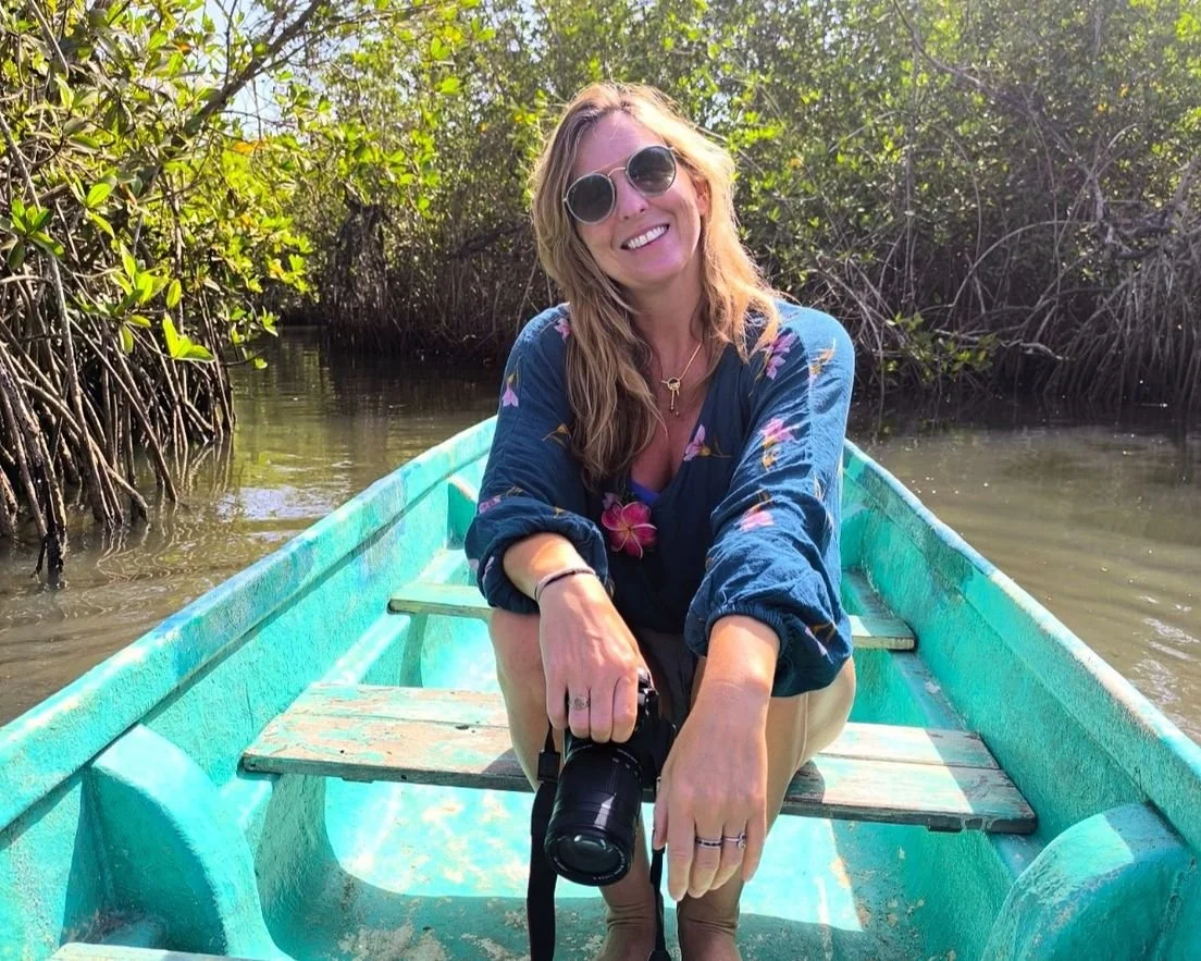 A woman with sunglasses and long hair is sitting in a small boat on a narrow waterway surrounded by dense green foliage. She is smiling and holding a camera.