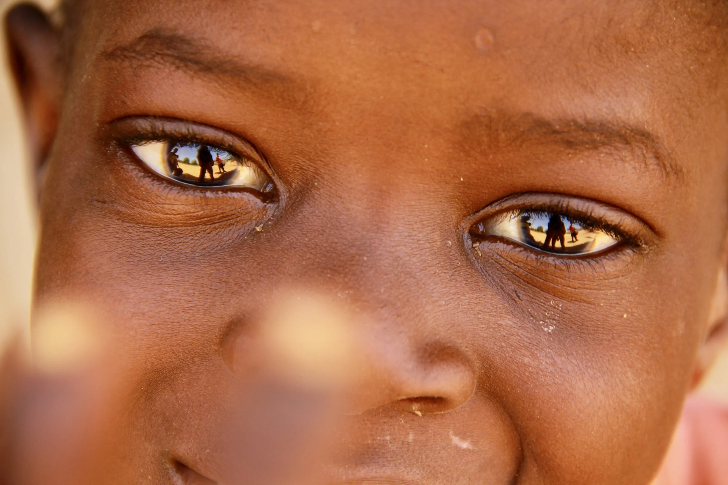 Close-up of a child's face showing their eyes reflecting a person and children outside in a sunny environment.
