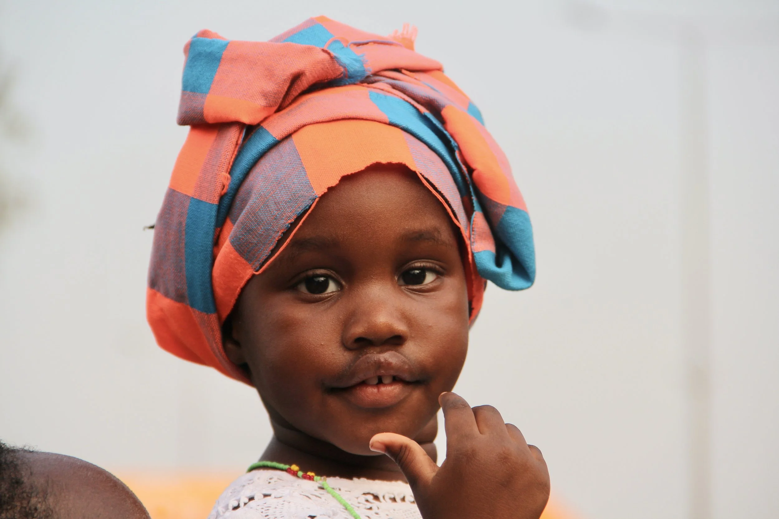 A young girl wearing a colorful headwrap and a white lace top, touching her chin with her finger and looking at the camera.