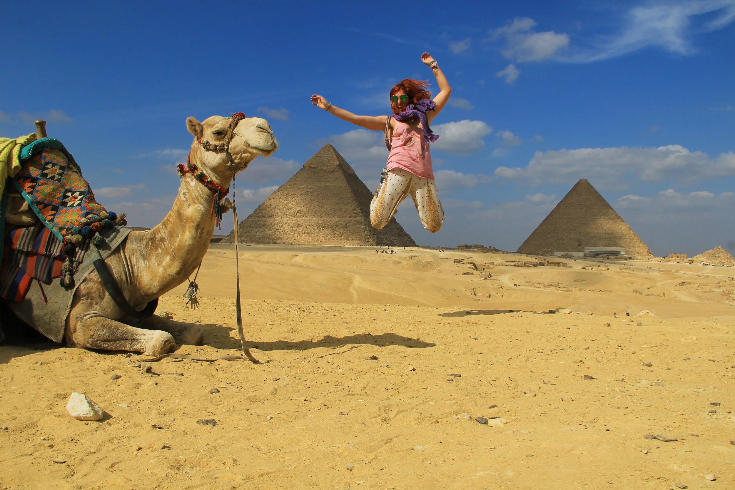 A woman jumping in front of the Pyramids of Giza with a camel sitting nearby on sandy terrain under a blue sky.