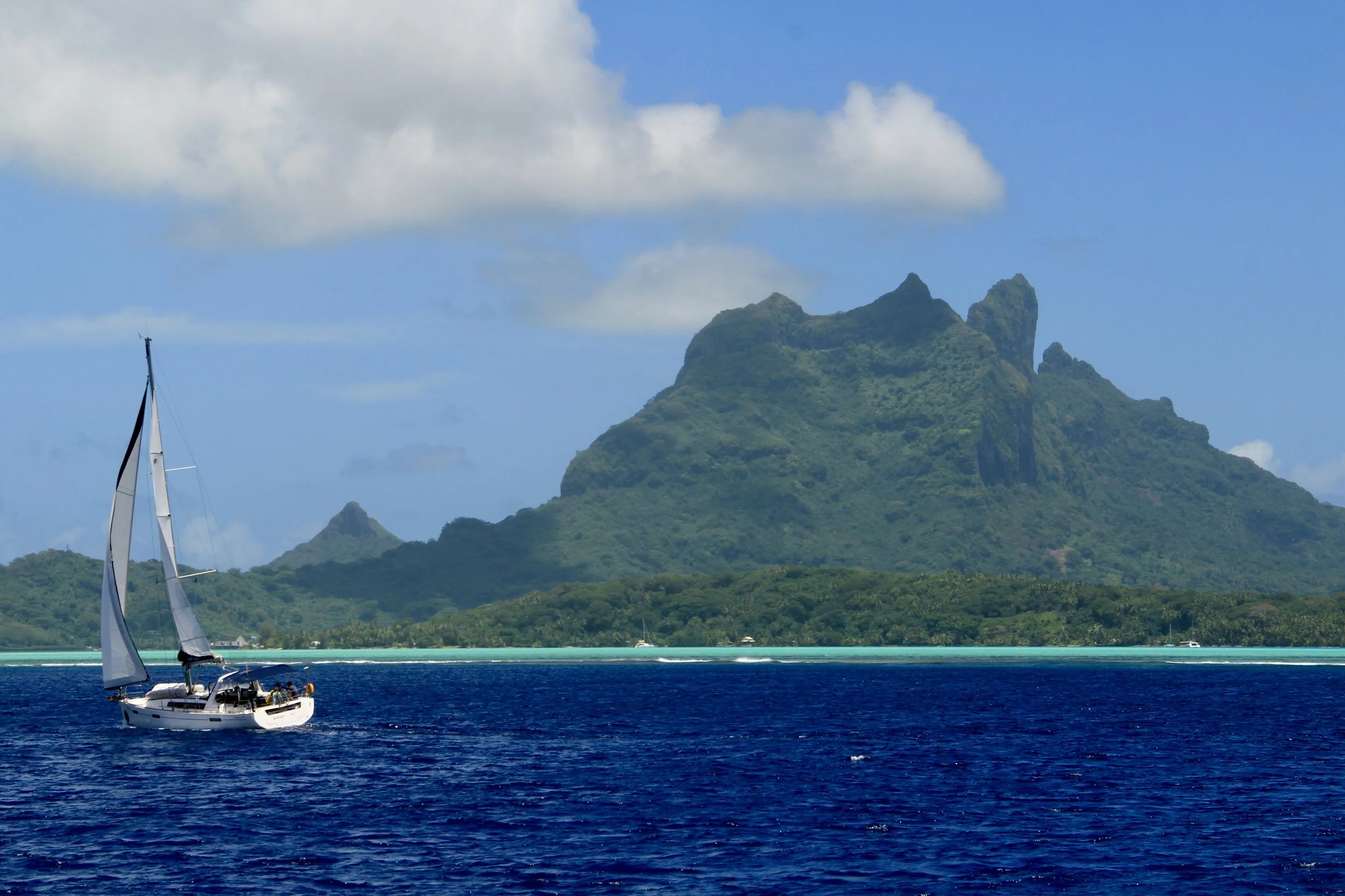 A sailboat with white sails on deep blue water in front of Green lush island with mountainous terrain, under a partly cloudy sky.