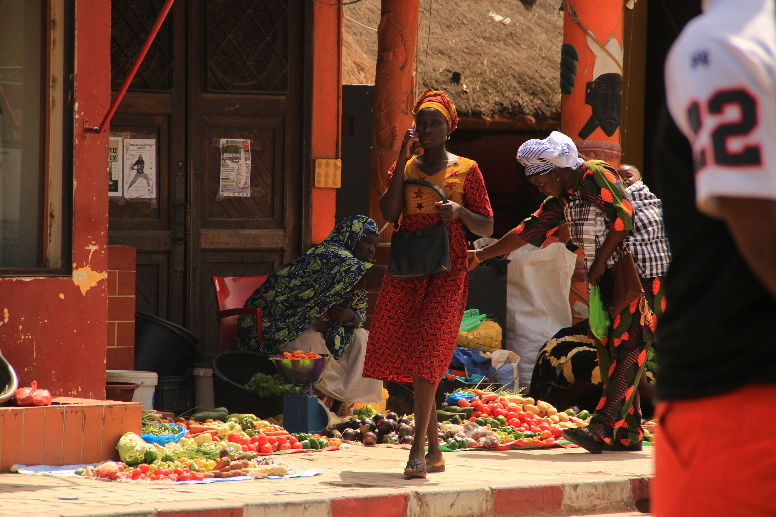 Women selling vegetables at an outdoor market in a vibrant, colorful setting with traditional attire and a wooden door in the background.