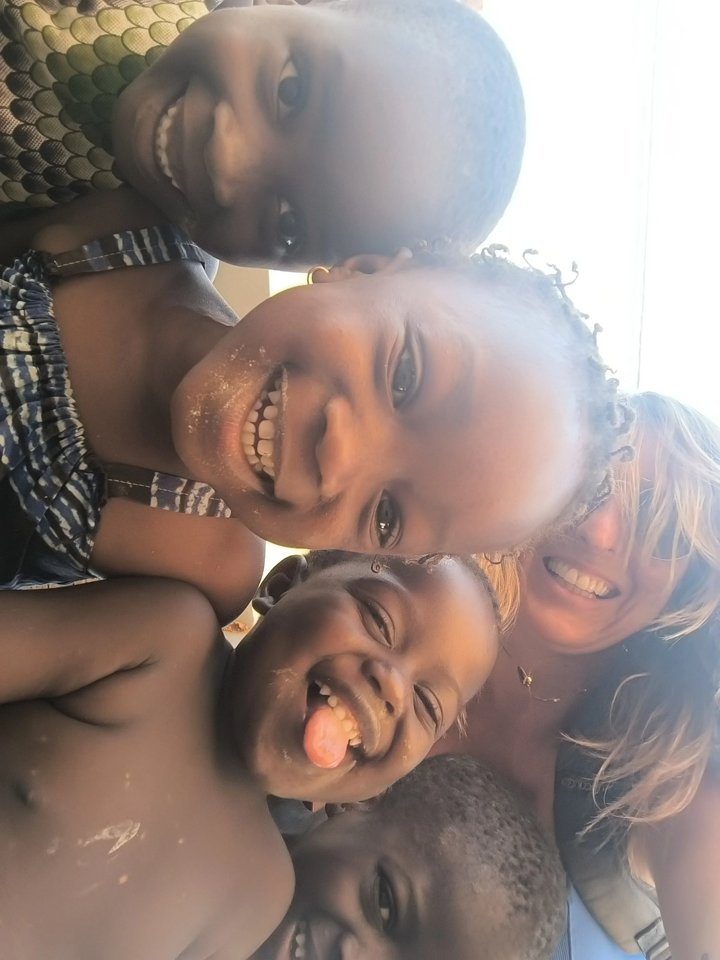 A group of five smiling children and a woman taking a selfie outdoors.