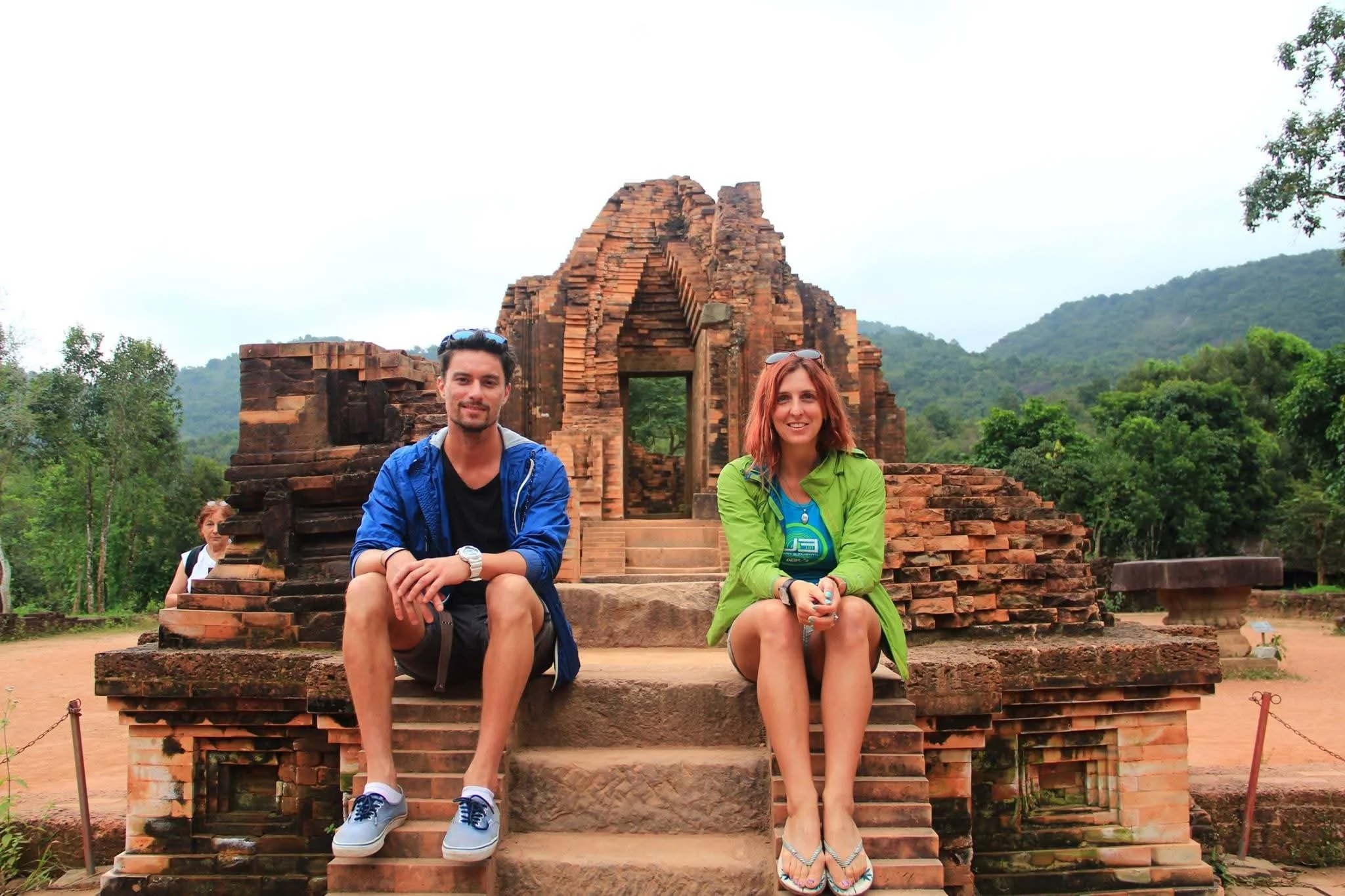 A man and a woman sitting on steps in front of ancient brick ruins, surrounded by green trees and hills.