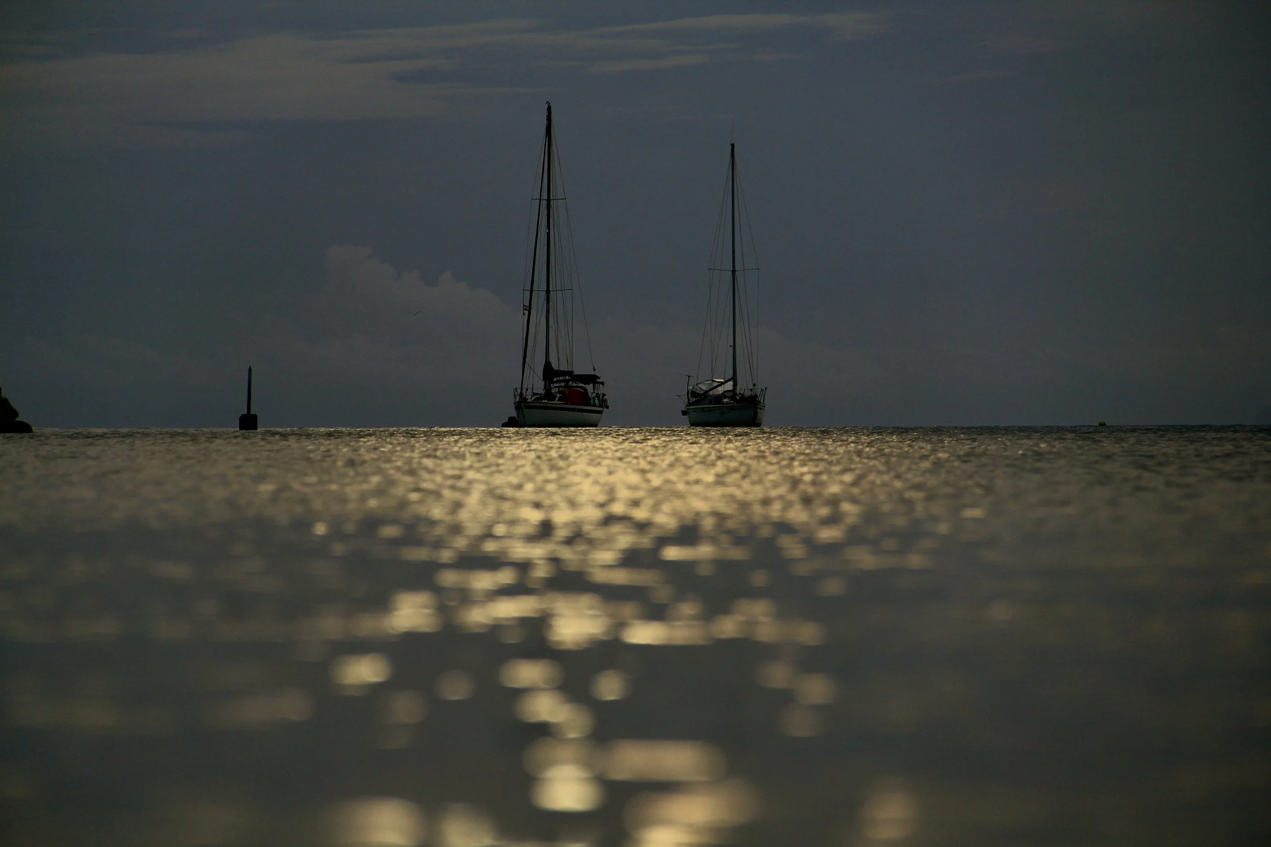 Two sailboats floating on calm water during a sunset or sunrise, with reflections shimmering on the surface.