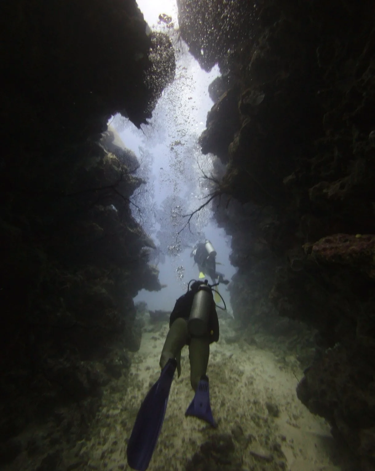 Underwater photo of two scuba divers exploring a narrow underwater canyon with rocky walls and bubbles rising to the surface.