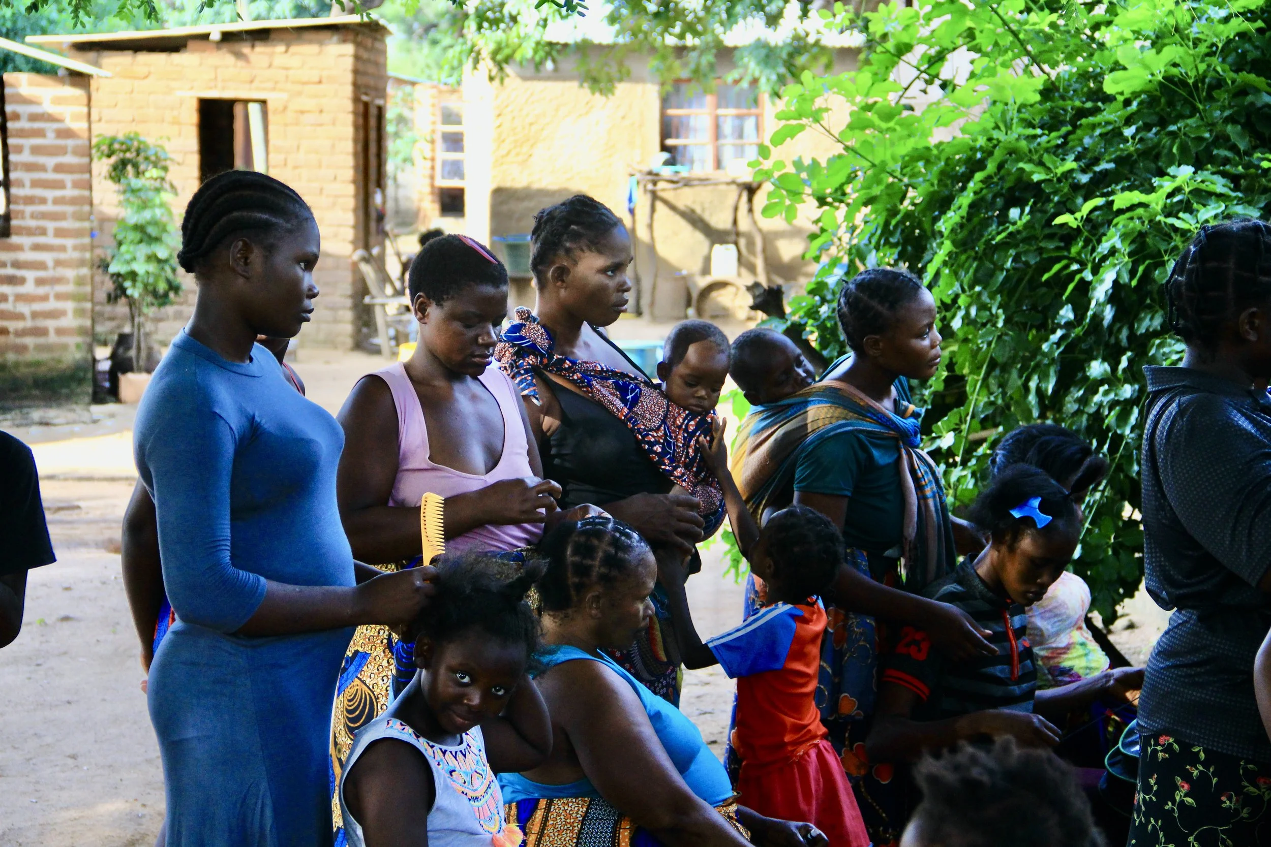 Group of African women and children gathered outdoors near green trees, with some sitting and others standing, in a rural setting with brick houses in the background.