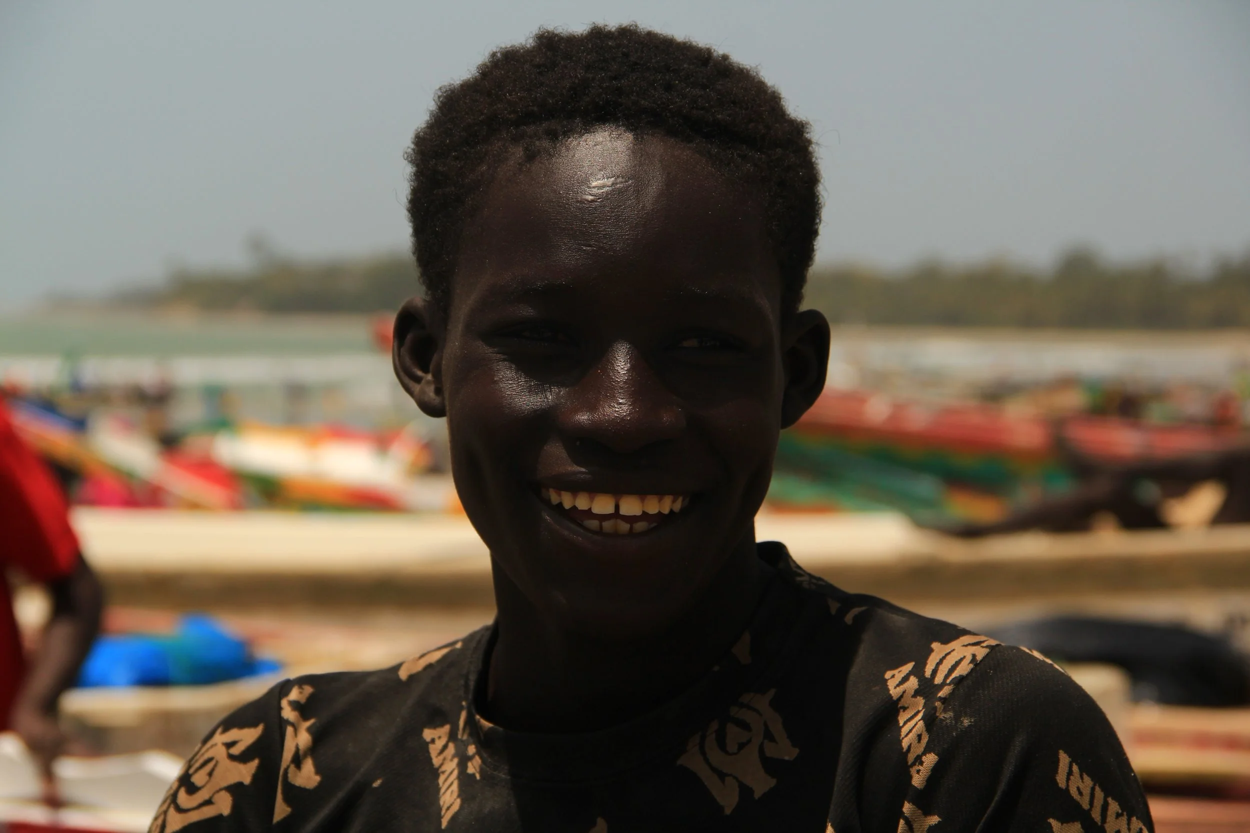 A smiling young man with dark skin and short curly hair, wearing a black shirt with gold patterns, standing outdoors near a beach with boats in the background.