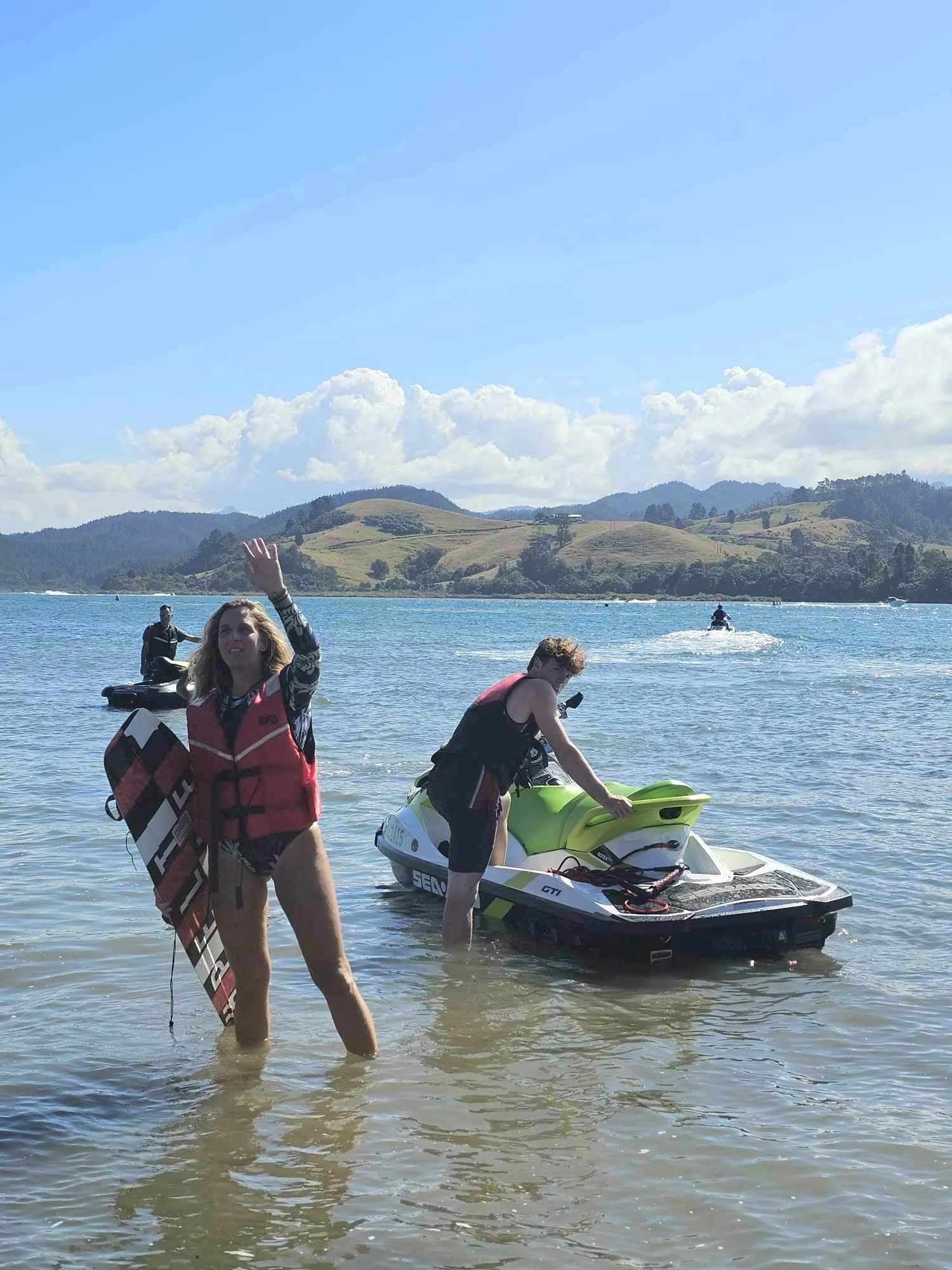 Two people in life jackets by jet skis on a beach with mountains and blue sky in the background.