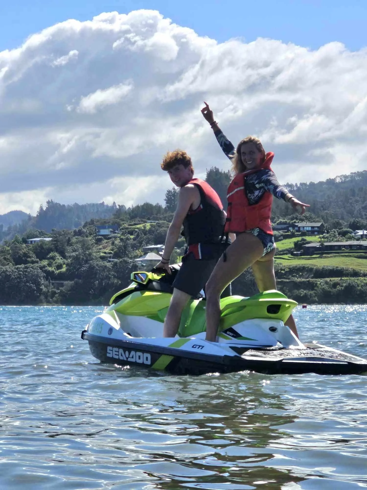 Two people on a green and white Sea-Doo jet ski on a body of water, with hilly landscape and houses in the background, under partly cloudy skies.