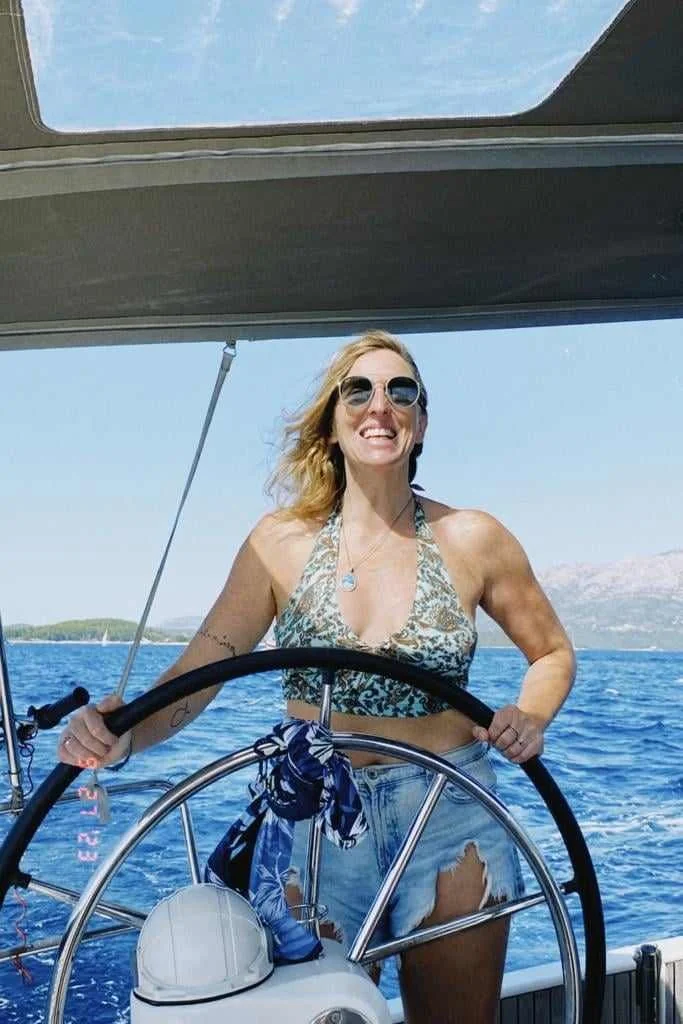 Woman in sunglasses and a bikini top steering a sailboat on the water during daytime with a scenic background.