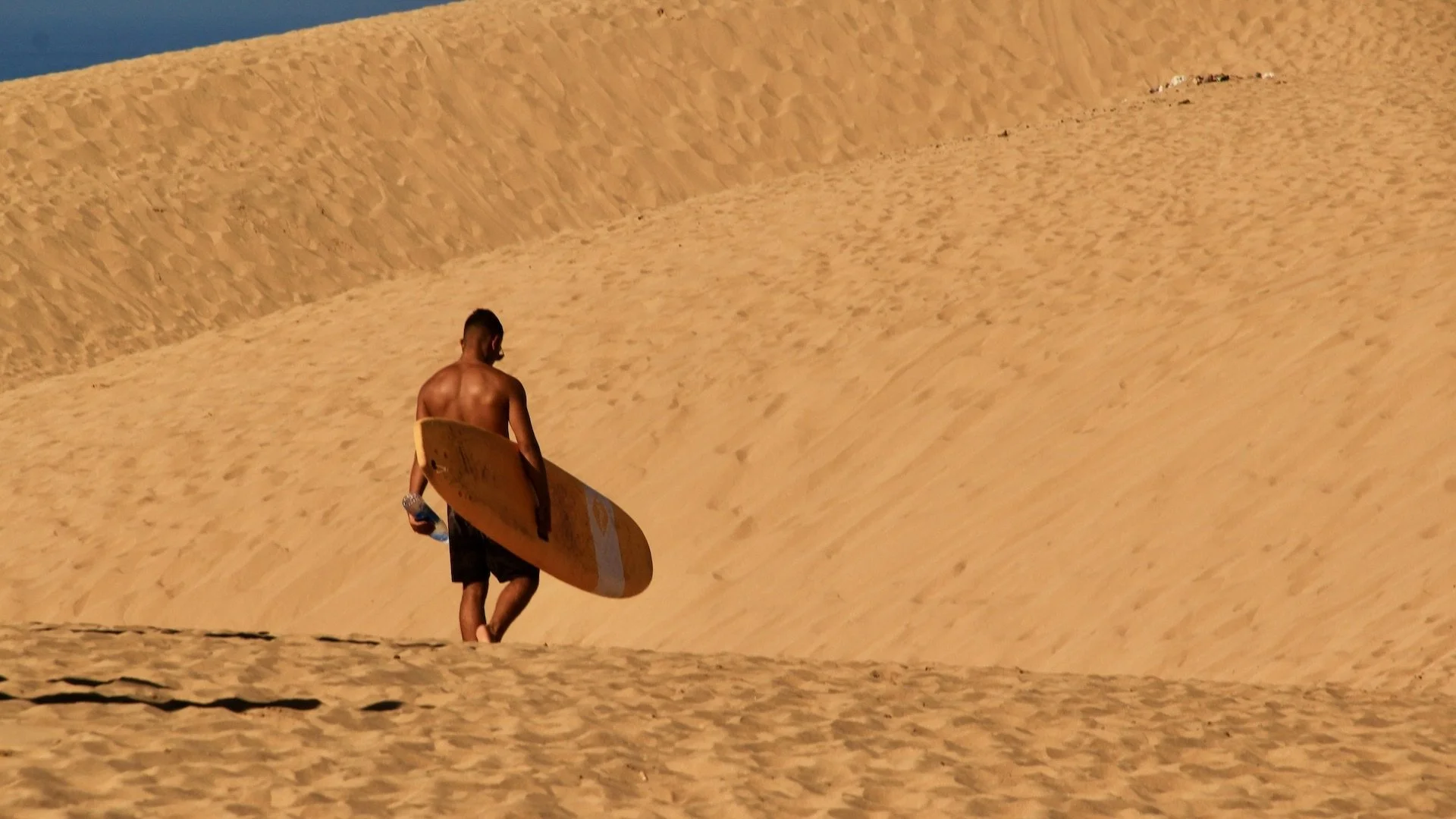 A shirtless man walking on the sand dunes, holding a yellow surfboard in one hand and a water bottle in the other, with a vast desert landscape in the background.