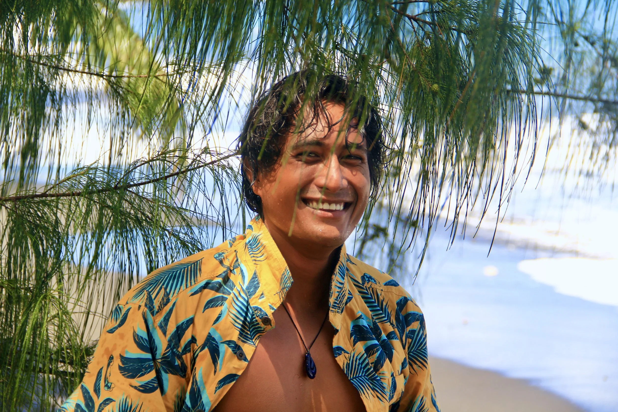 A smiling man with wet hair wearing a bright yellow Hawaiian shirt with blue leaves, standing on a beach with water and trees in the background.