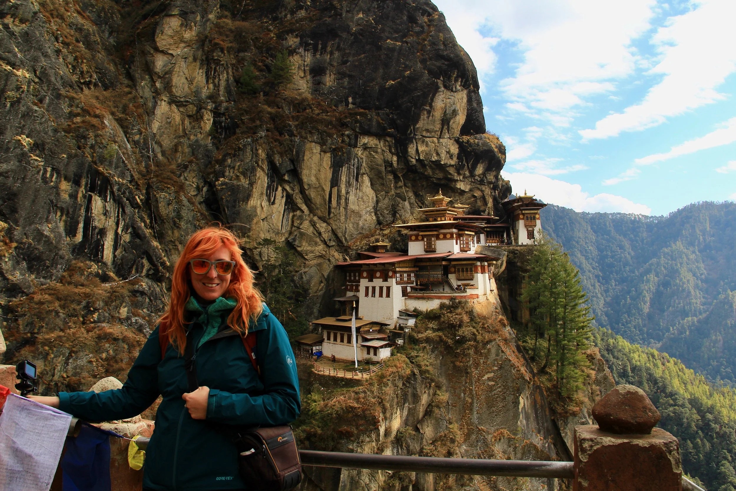 A woman with red hair and sunglasses smiling at a mountain monastery in Bhutan, with rocky cliffs and green trees in the background.