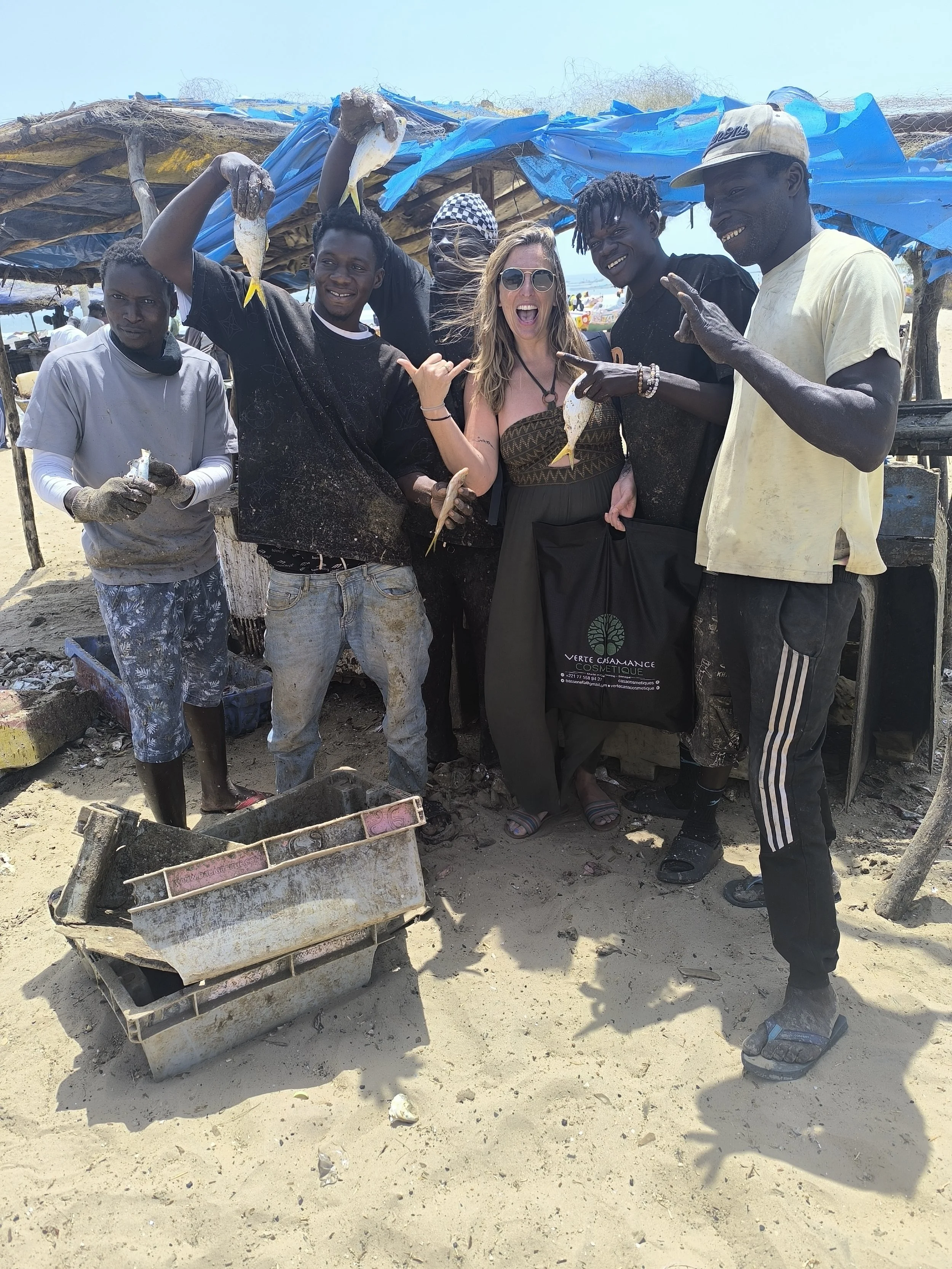 Group of people, including a woman with sunglasses, smiling, holding fish, on a sandy beach, with makeshift blue tarp shelter behind them.