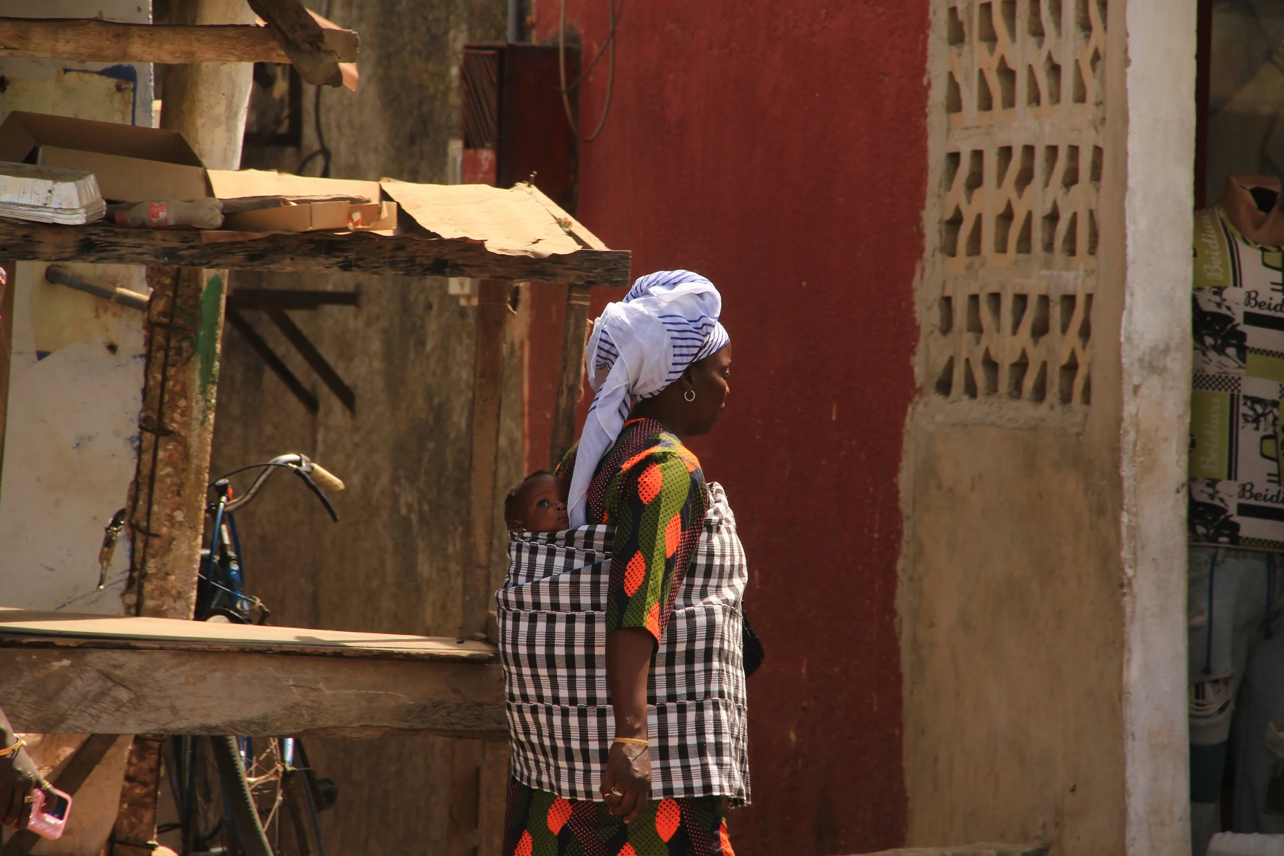 A woman wearing a colorful dress and headscarf carries a baby in a checkered sling on her back, walking past a wooden stall with a bicycle nearby, against a background of weathered buildings with a lattice brick wall.