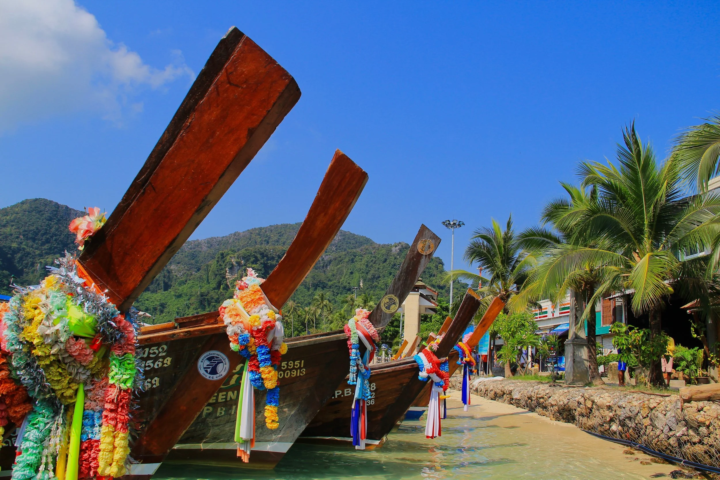Traditional wooden longtail boats decorated with colorful flowers and ribbons docked along a tropical shoreline with palm trees and hills in the background.