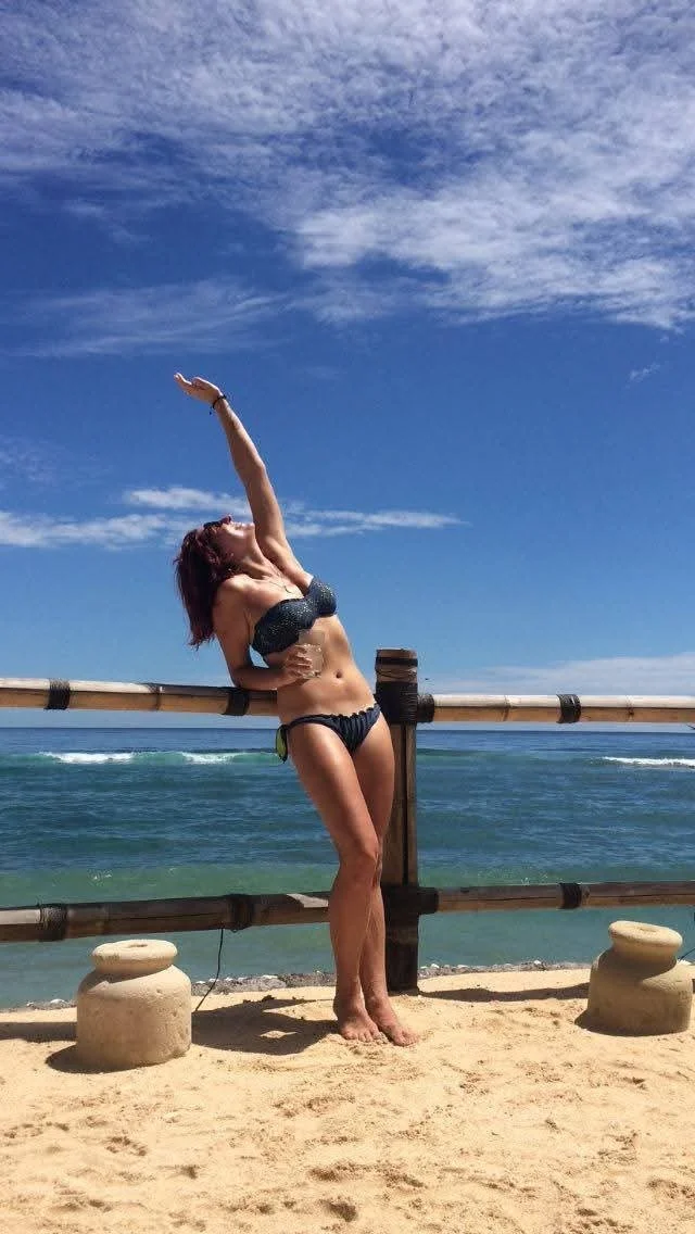 Woman in a black bikini standing on a sandy beach next to a wooden fence, with the ocean and a blue sky with clouds in the background, stretching her arm upward.