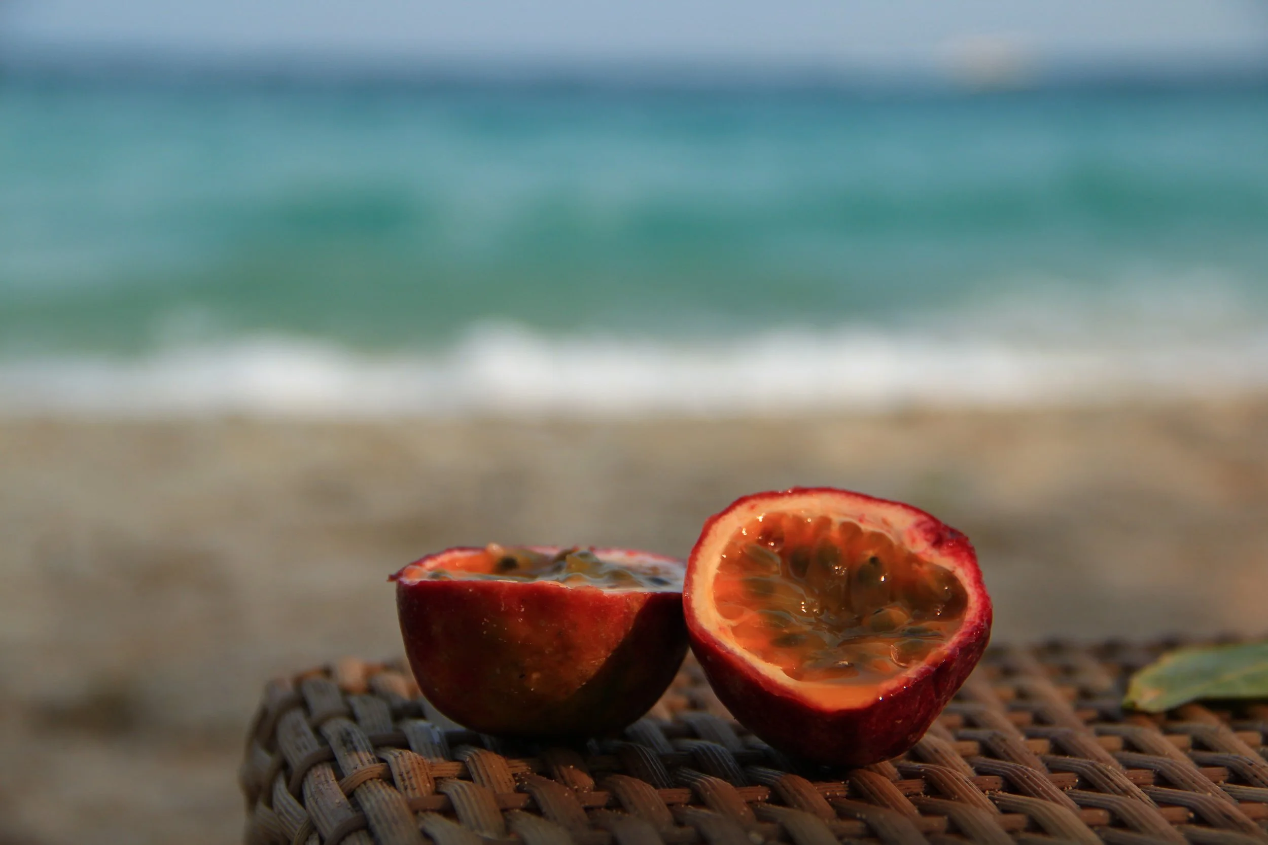 Half and a whole passion fruit on a woven surface with blurred ocean waves in the background.