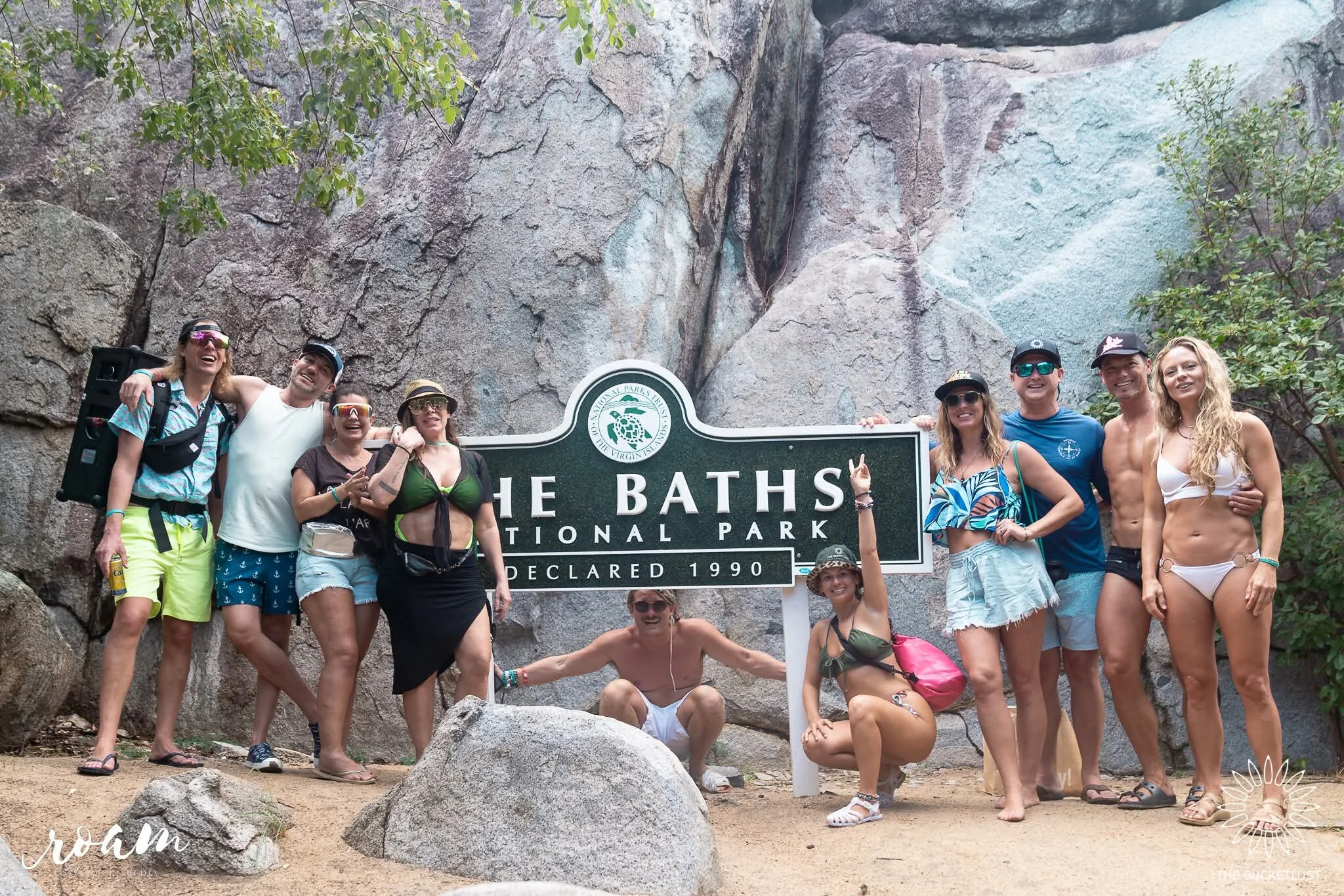 A group of people in swimsuits and summer attire standing in front of a sign at The Baths, a national park, with large rock formations in the background. They are smiling and posing for a photo.