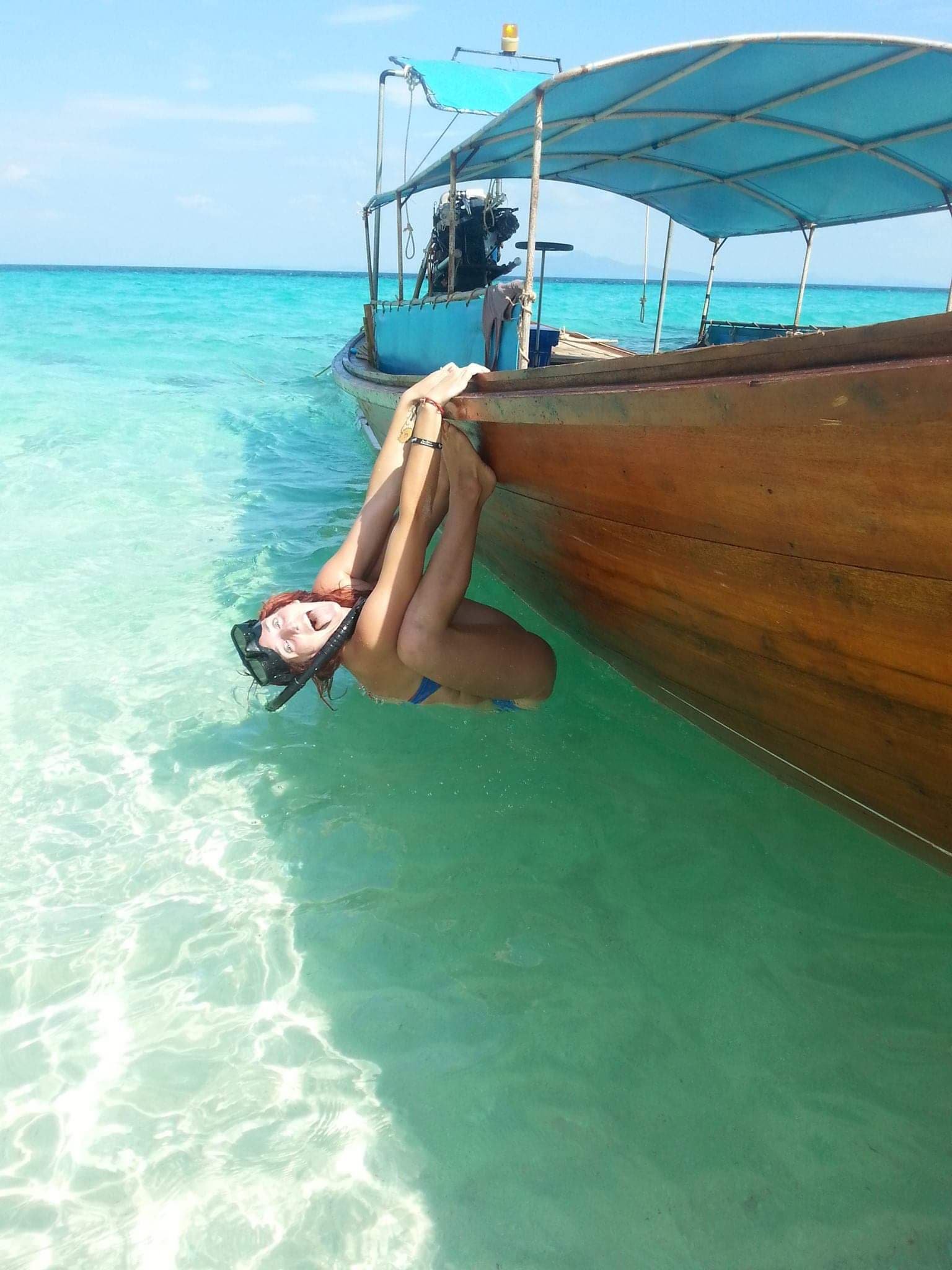 A woman clinging to the underside of a boat near the water, holding onto the boat's side with her arms, in a tropical setting with turquoise water and a clear sky.